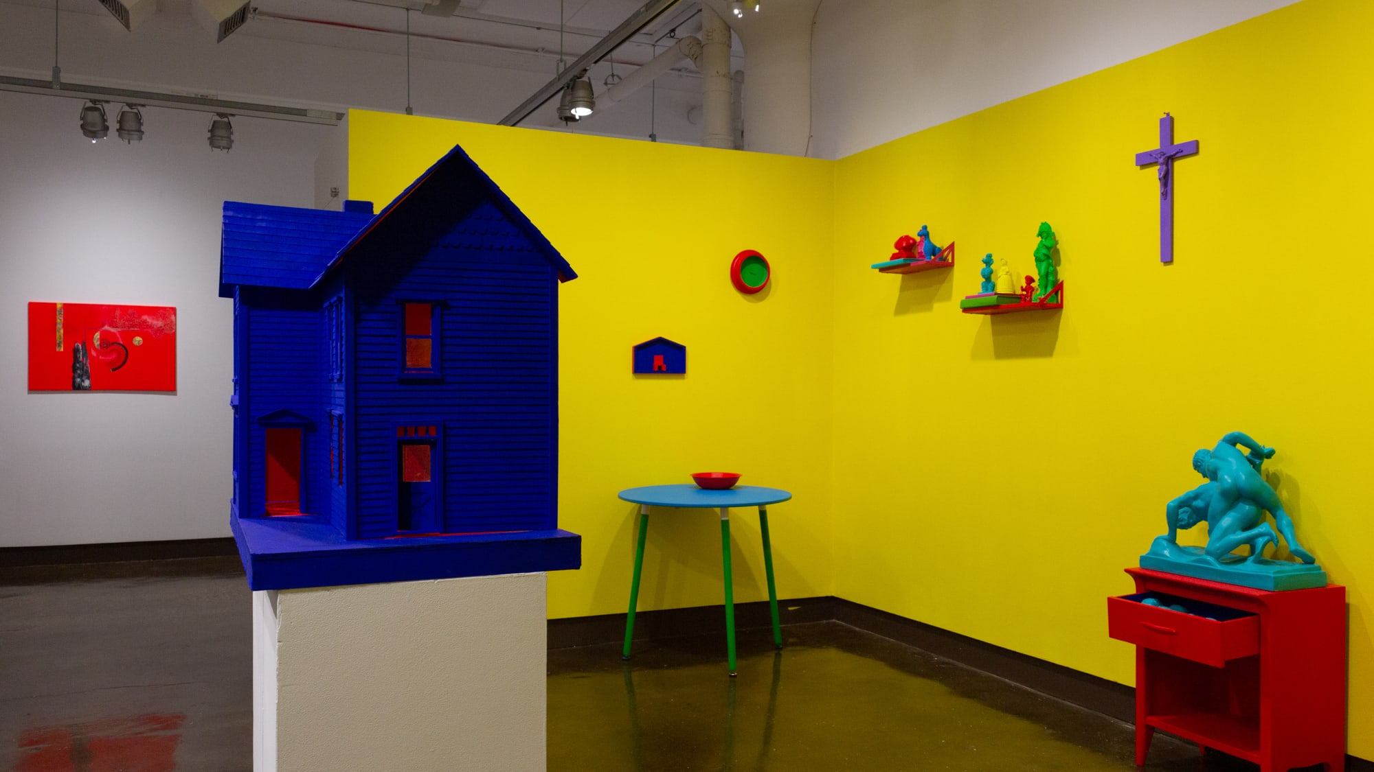 On the right is an inset space, painted yellow, with hyper-pigmented painted objects common to a household including a nightstand, chair, and large dollhouse. On a nearby wall to the left is a bright red abstract painting.