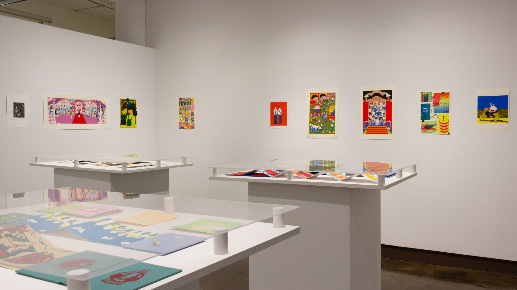 In the corner of an inset space are multiple colorful prints in a horizontal line across two adjacent walls. In the center of the space are three white pedestals, each containing multiple printed books, with plexiglass each of the three surfaces.