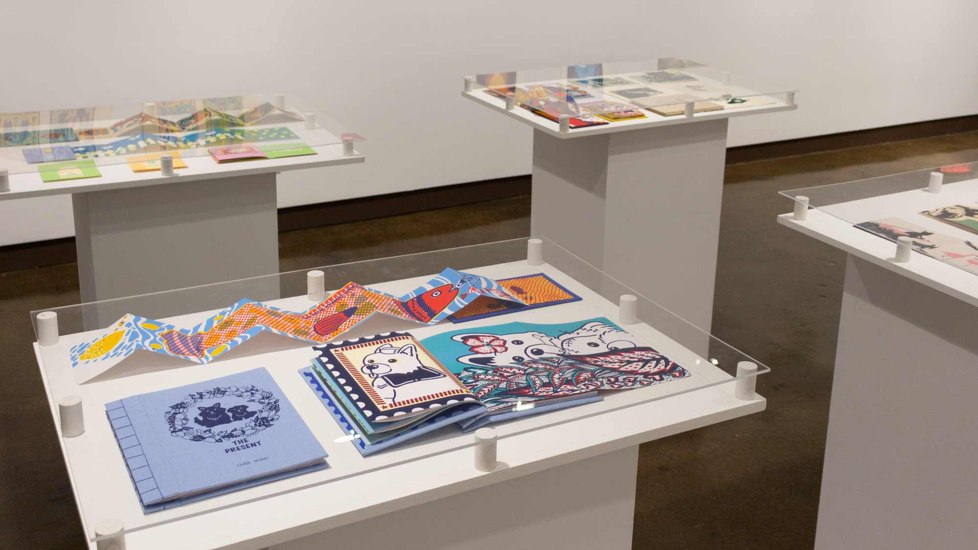 Four white pedestals, each containing multiple printed books, and covered in large sheets of clear plexiglass.