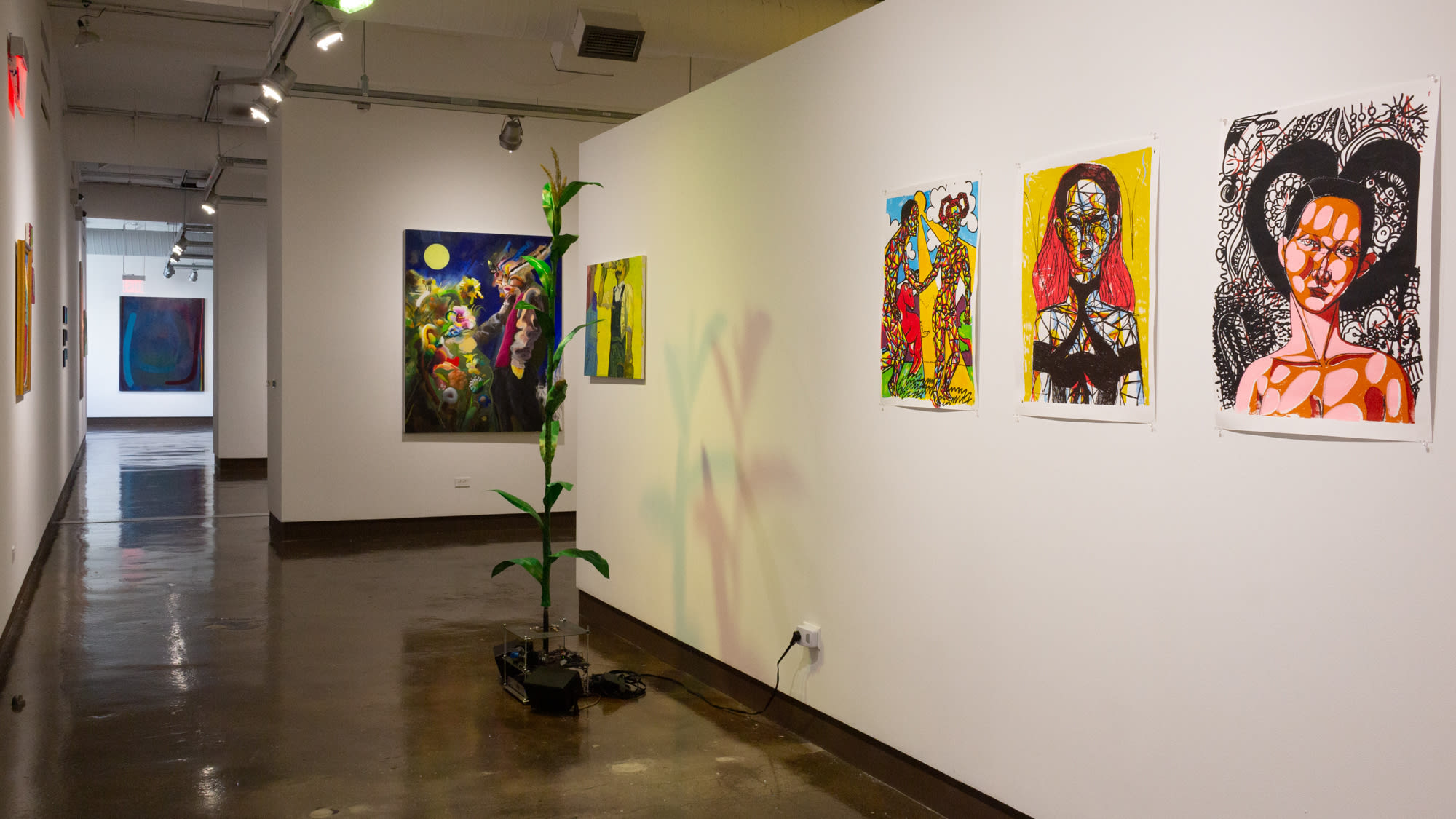 On the right are several paintings hung on a white wall, and in the center of the frame is a tall plastic cornstalk connected to a series of electronic motors and sensors. On the left is a view of a hallway leading to adjacent gallery spaces.