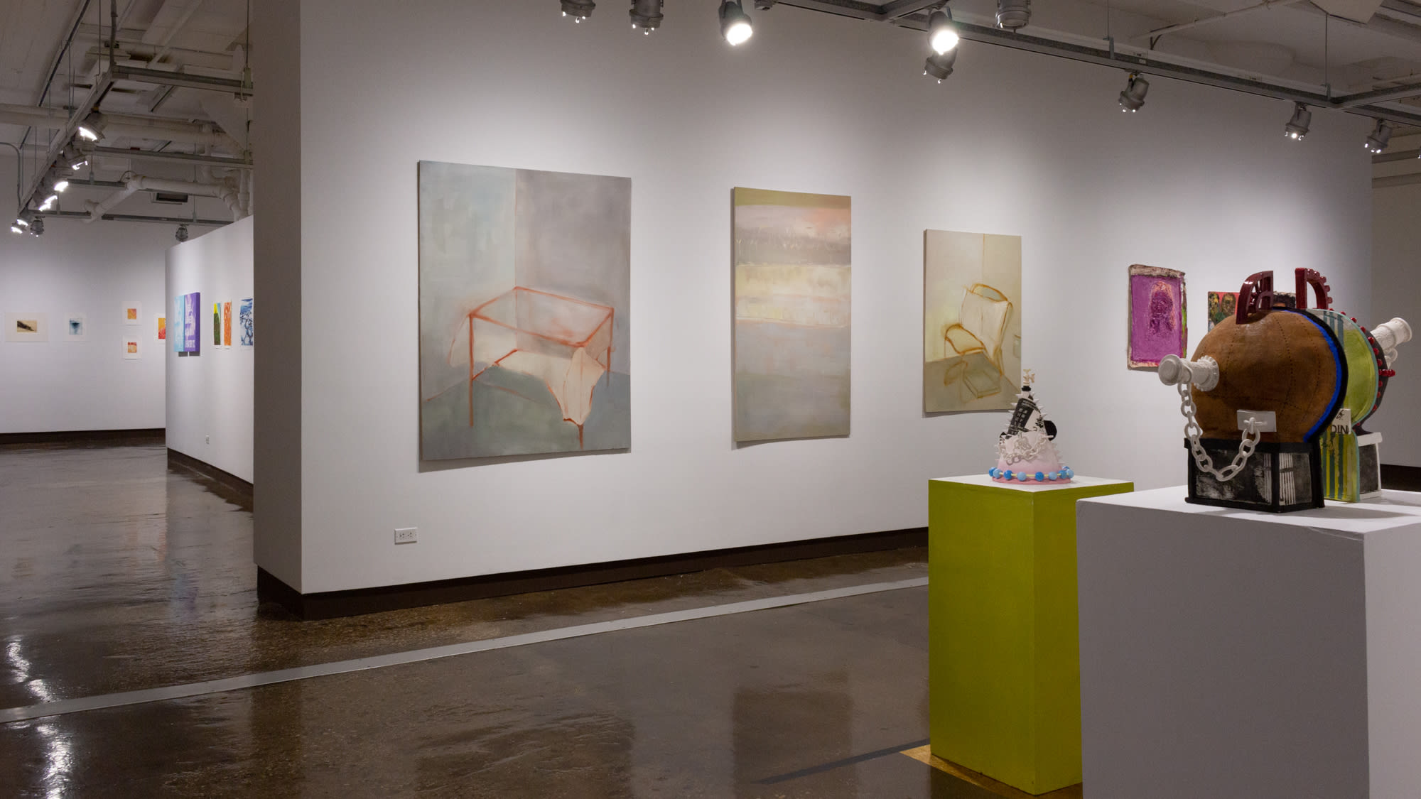 On the left are multiple colorful prints on two walls, on the right is another wall with three subtly colored paintings depicting very minimal spaces with the armatures of furniture items rendered in lines. On the lower left corner of the image are two sculptures in geometric shapes, each on a pedestal.