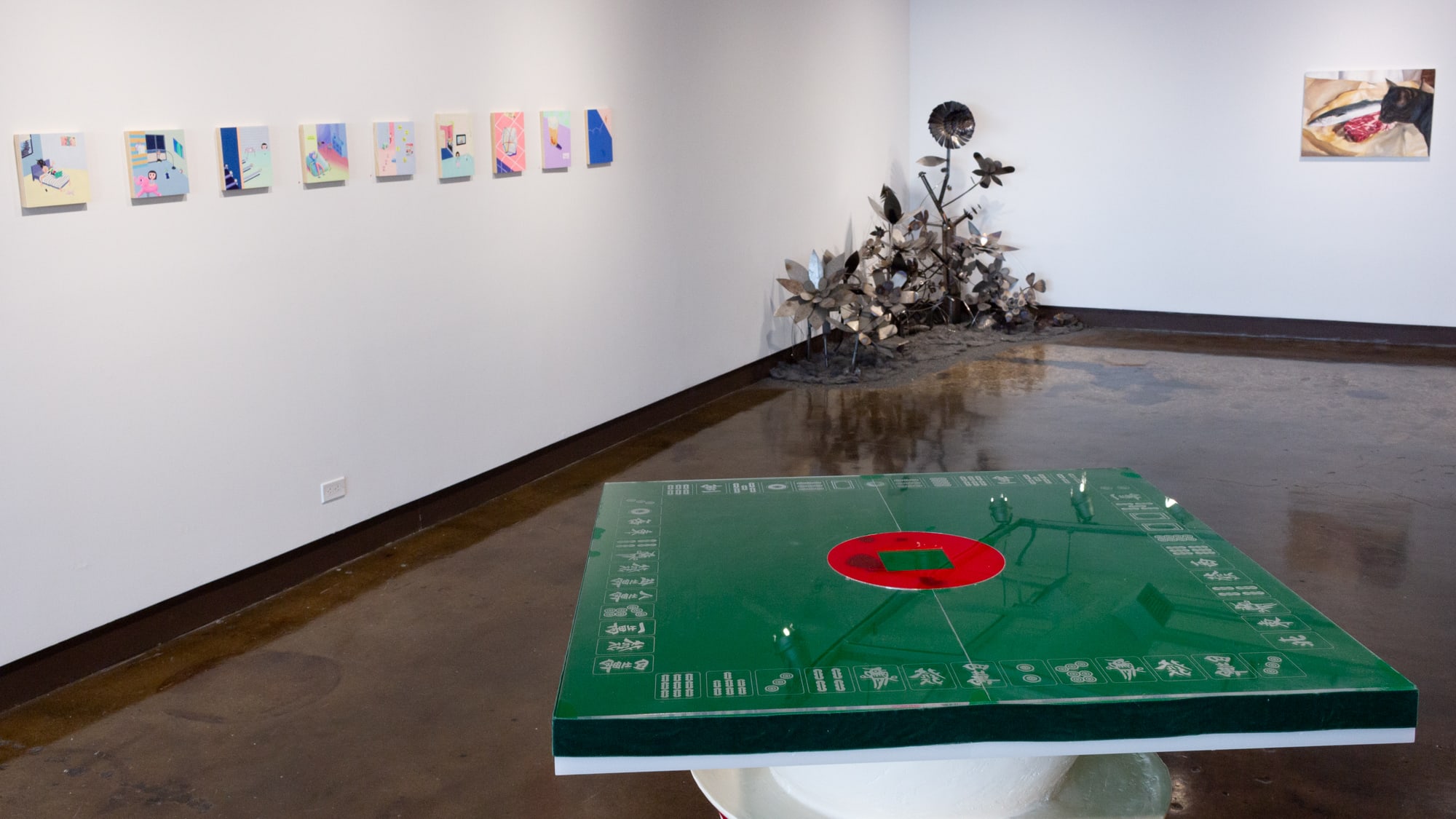 Photograph from front gallery space of nine small pastel-colored paintings hung in a horizontal row, in the center of the frame is a green and red square surface, beyond it is a metal sculpture of a garden, and on the far right is a painting of a cat eying a large steak.