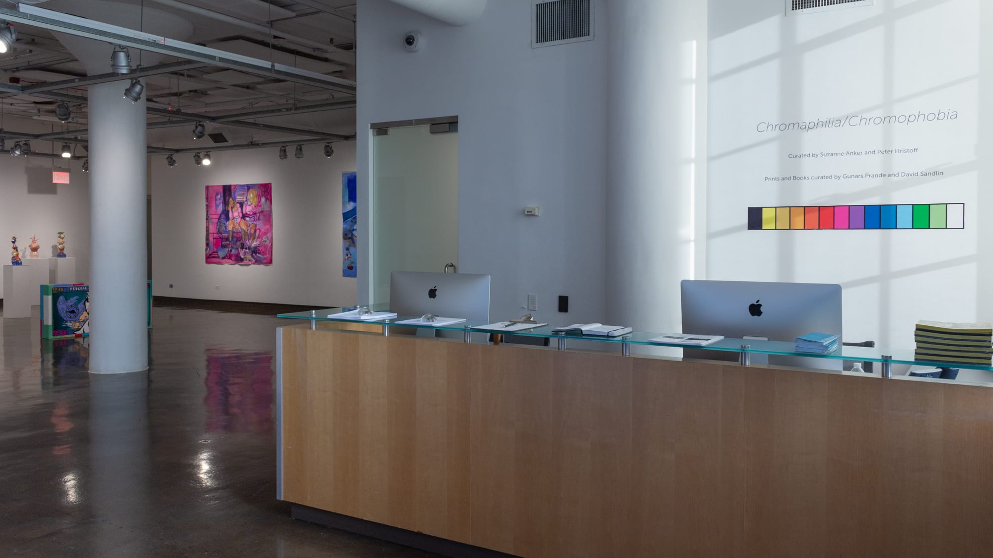 View from entrance of SVA Chelsea Gallery, with front desk to the right, the exhibition title "Chromophilia/Chromophobia" behind it on the wall, and to the left is a glimpse into the first gallery space.