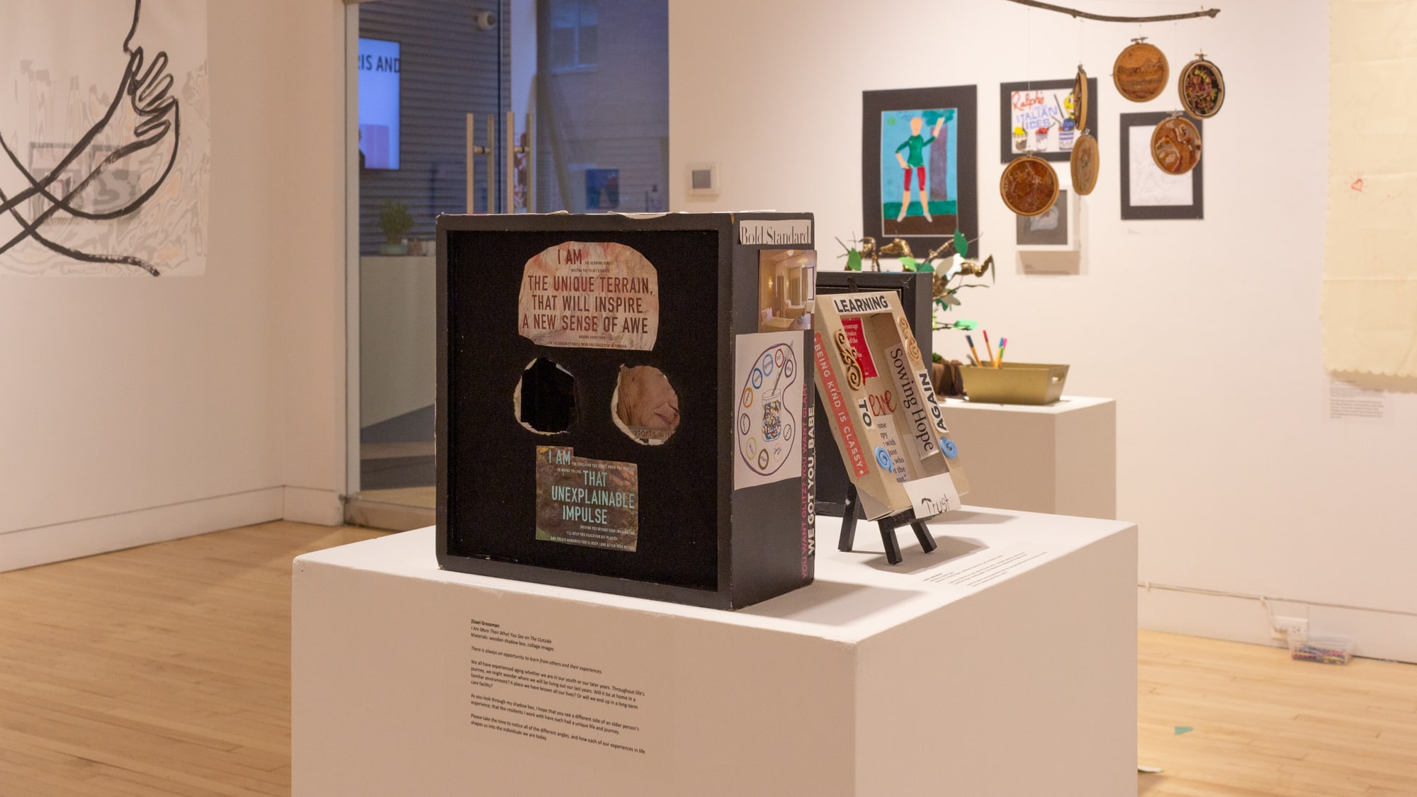 A variety of artworks by Art Therapy students against a white wall in gallery space. In the foreground are some small pieces on a square white pedestal.