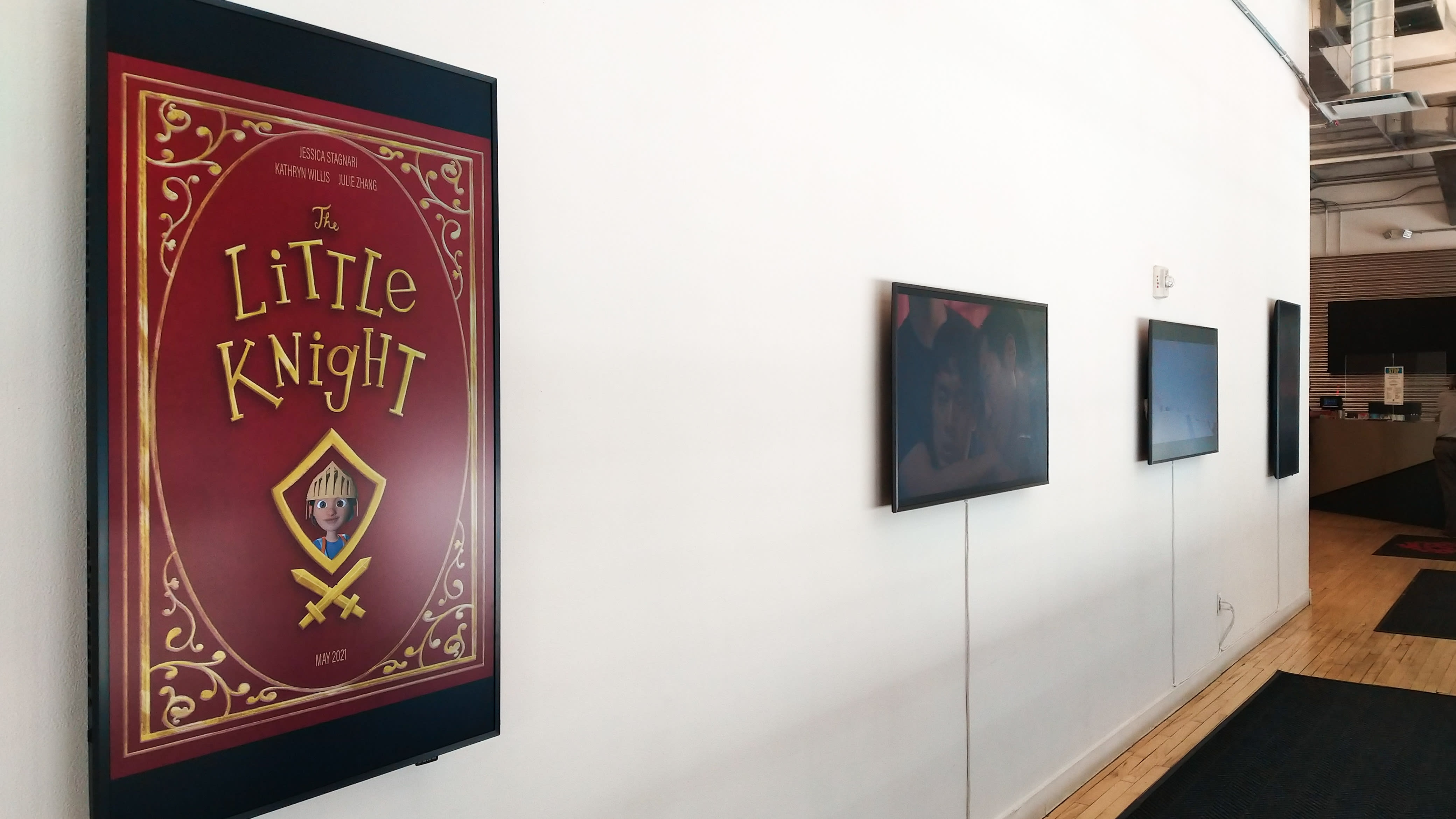 Four monitors on a white wall, the furthest to the left is vertical and shows a book cover for "The Little Knight" in red and gold.