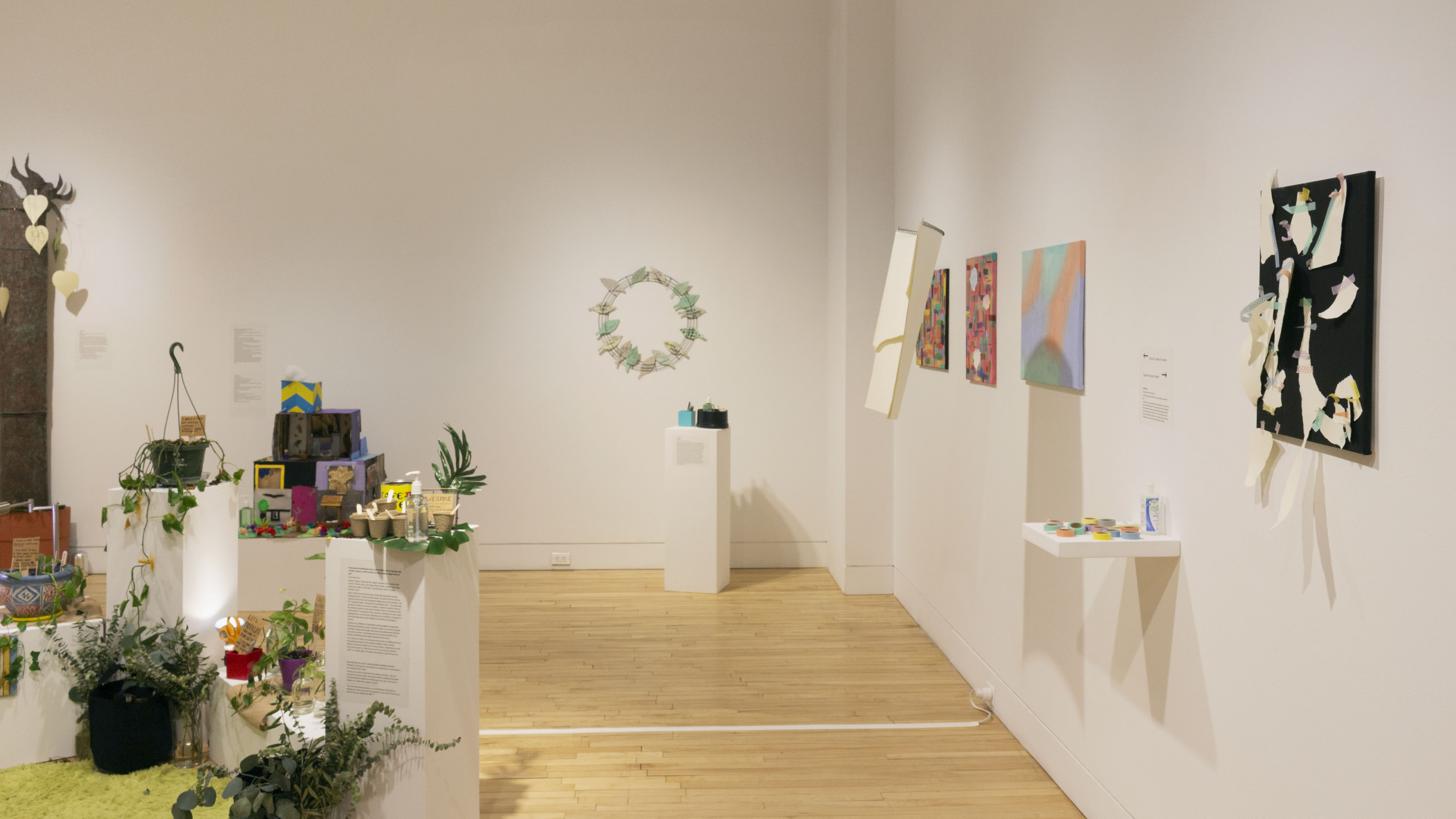 Multiple views of SVA Flatiron Gallery. There is a green rug in the center of the gallery with plants and soil around it. Paintings and artworks are hung on the walls. There are pedestals throughout the gallery with interactive pieces for visitors.