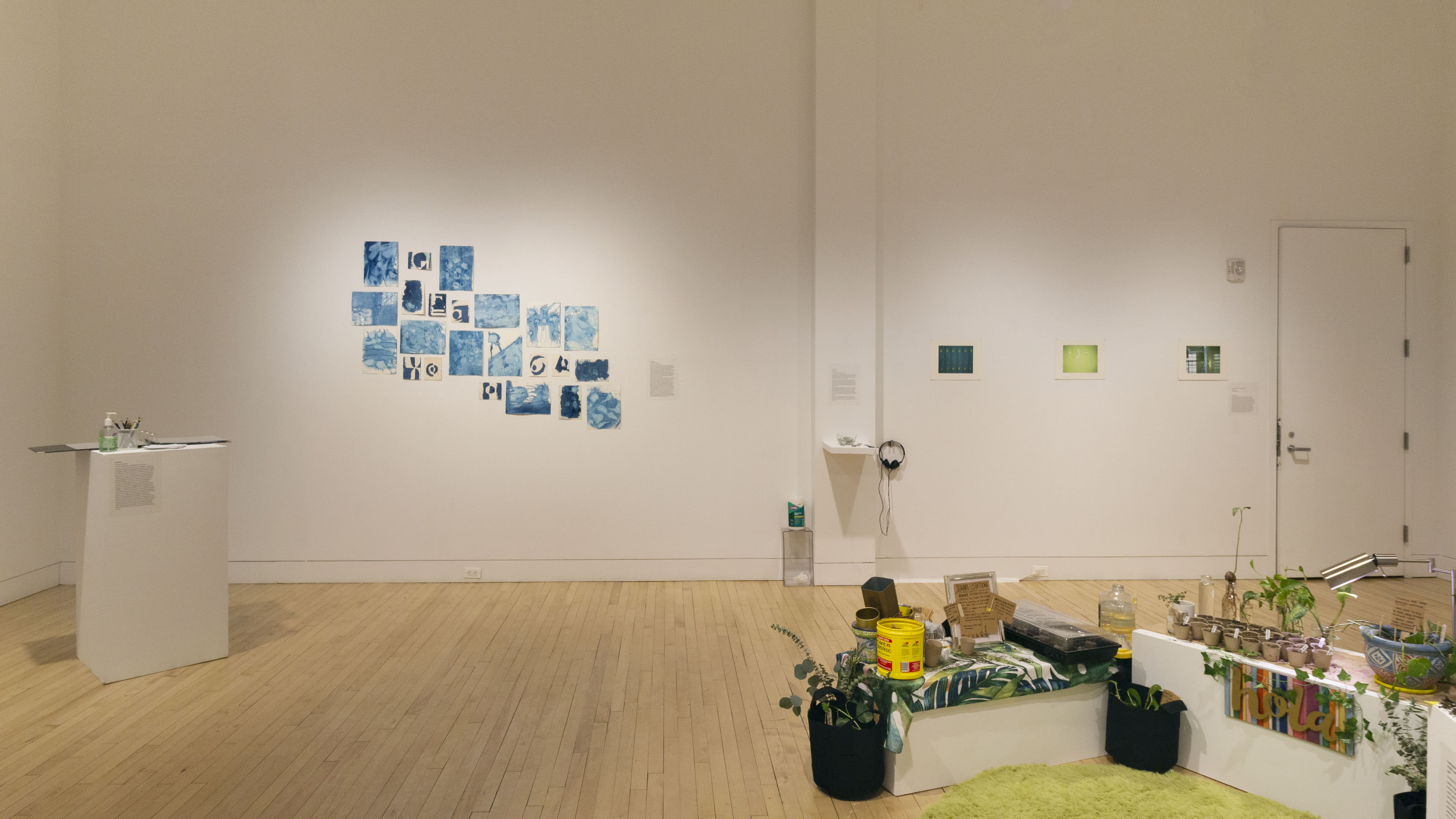 Multiple views of SVA Flatiron Gallery. There is a green rug in the center of the gallery with plants and soil around it. Paintings and artworks are hung on the walls. There are pedestals throughout the gallery with interactive pieces for visitors.