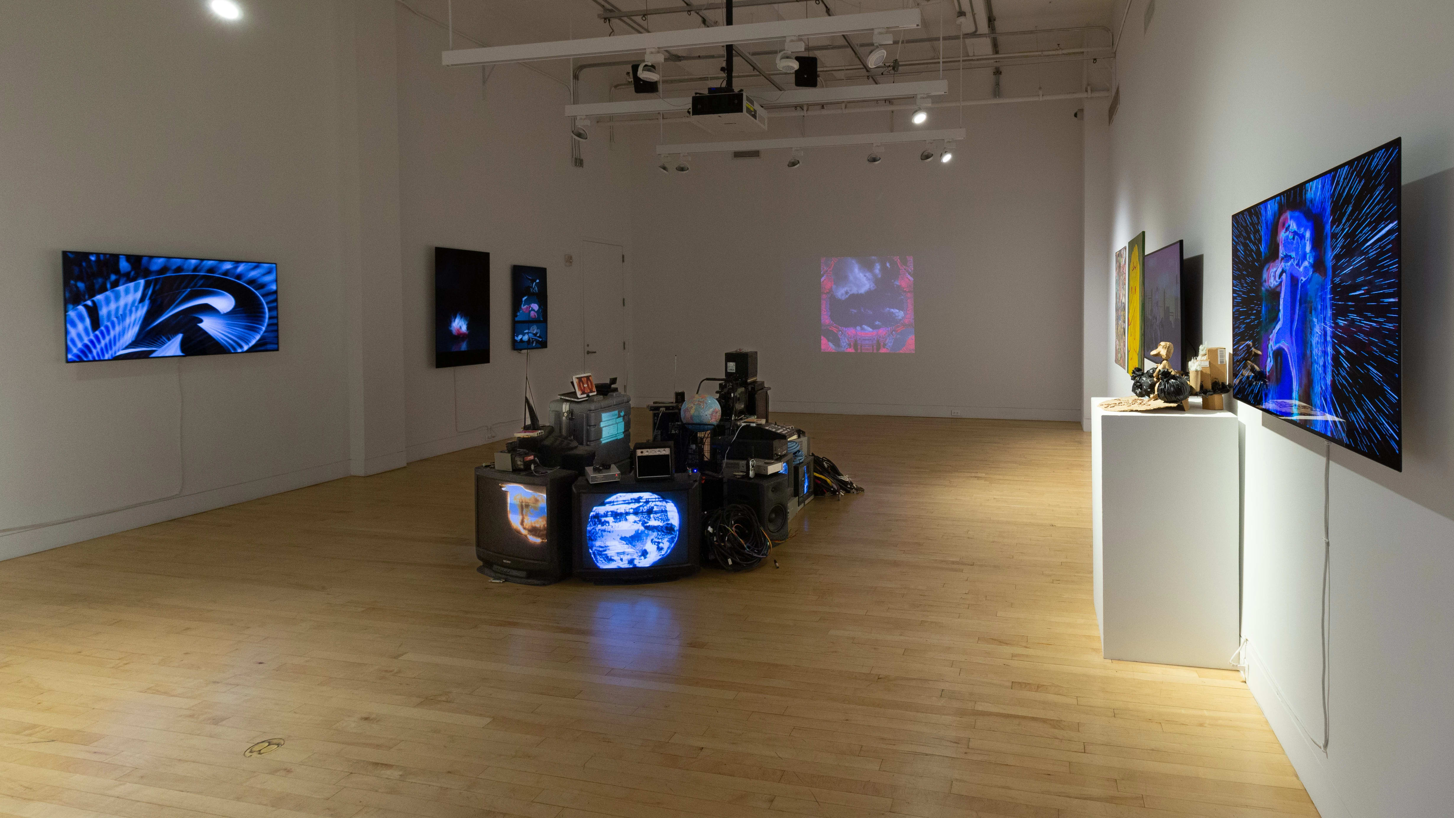 Inside the SVA Flatiron Gallery, the work of MFA Computer Arts students is on display. An installation of televisions, amplifiers, and other audio/visual equipment sits in the center of the room, while monitors and projections show art on the walls.