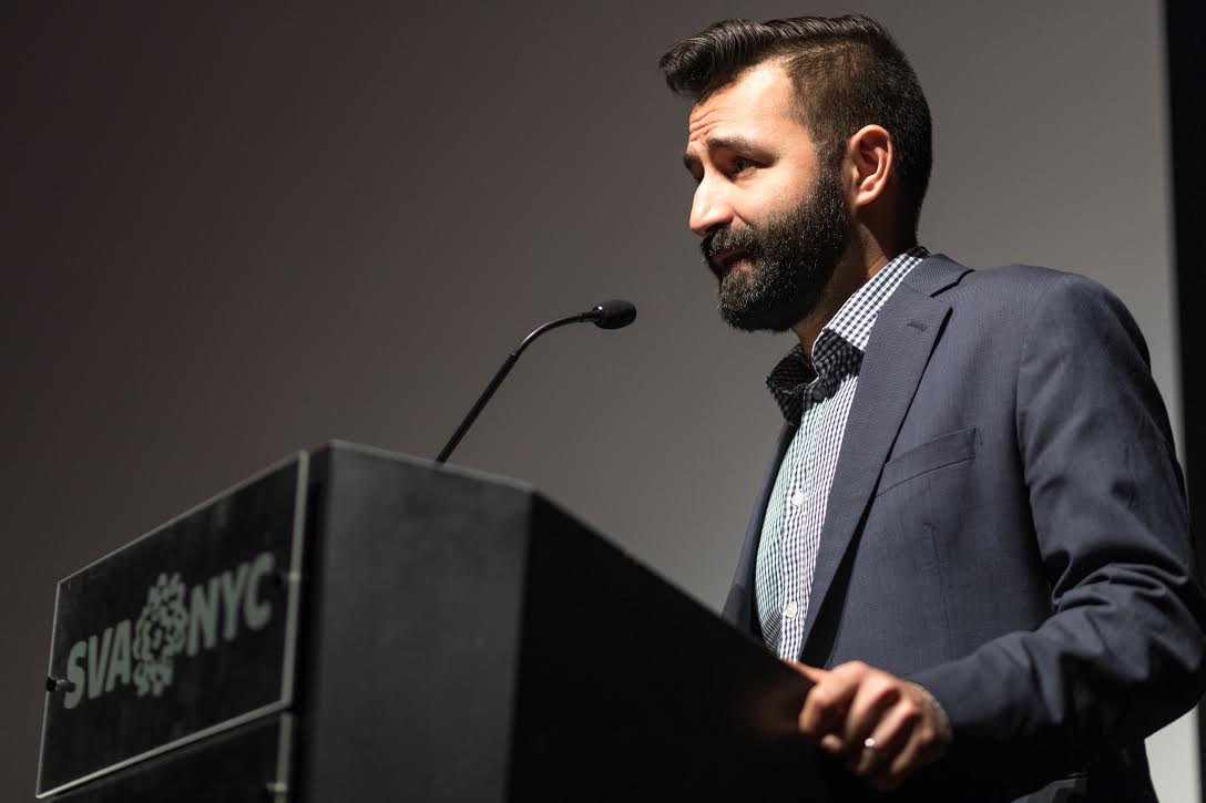 A man with a beard stands at a podium talking.