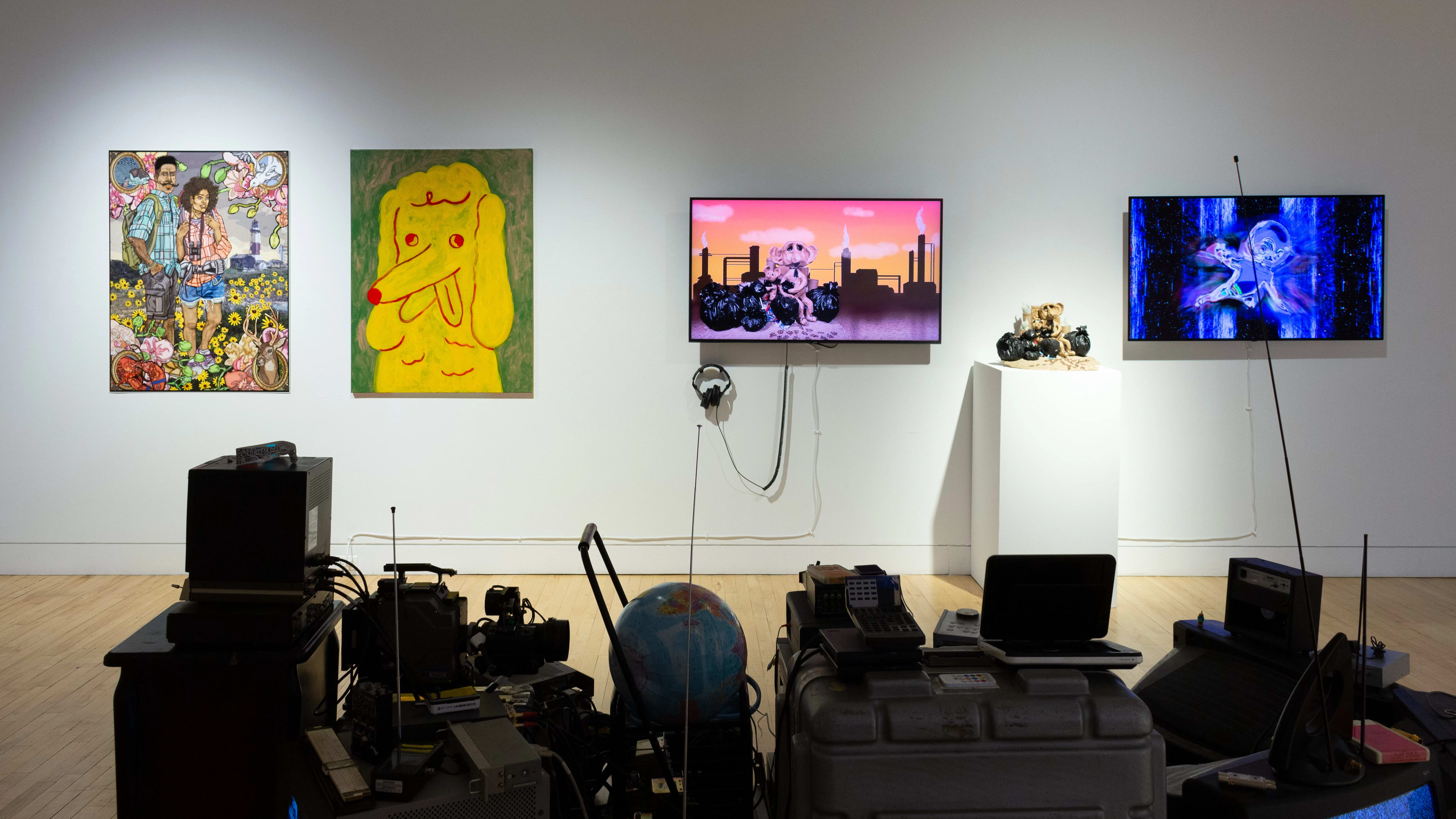 On the left side of a wall in the gallery, there are two paintings: one of a couple in hiking gear surrounded by flora and fauna, and one of a large yellow cross eyed dog with its tongue sticking out. The right side features two monitors showing video works, along with a sculpture on a pedestal that also appears on one of the monitors.