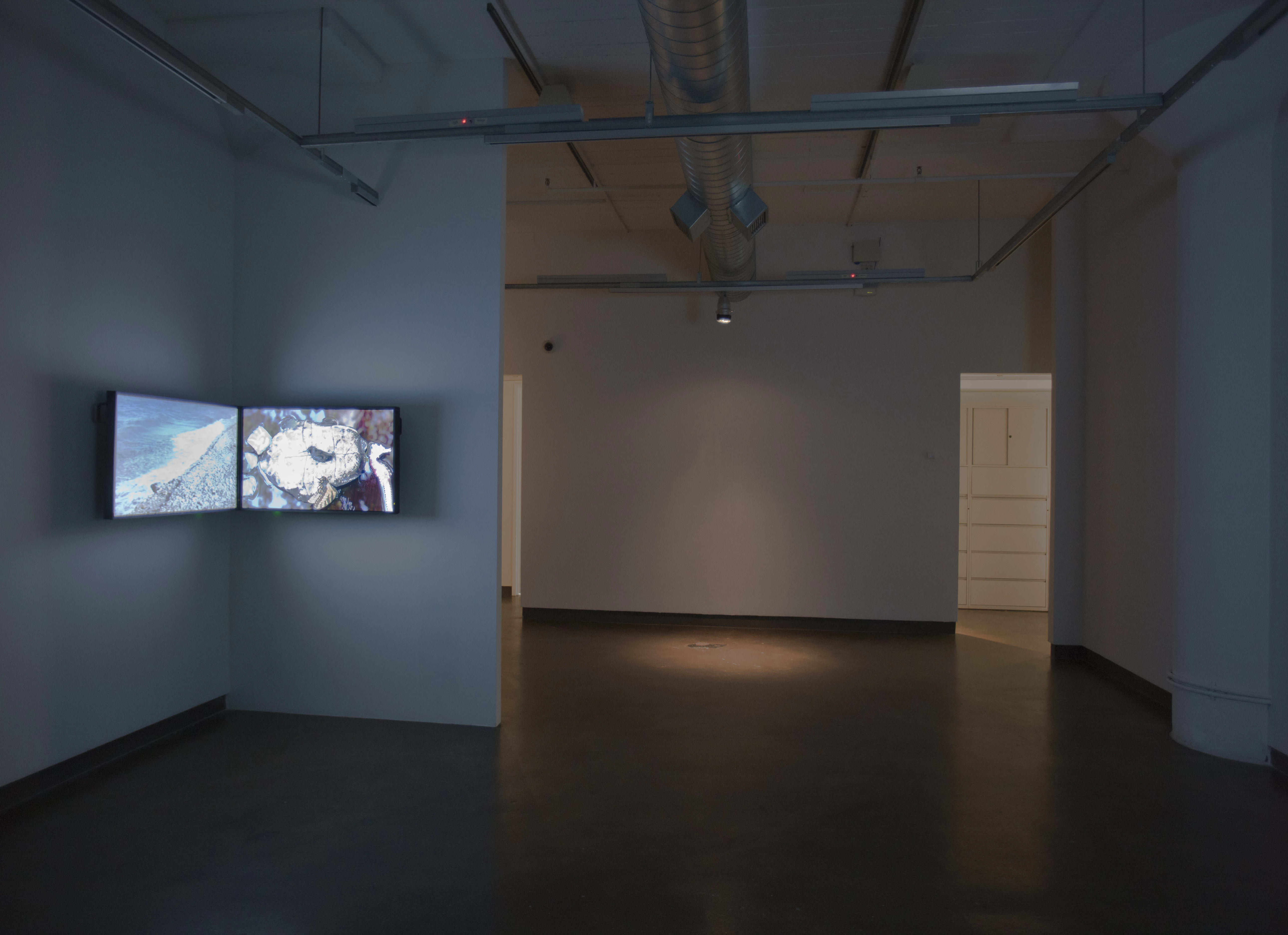A dark room that is connected to another room.  The first room contains two tv's side by side in the corner.