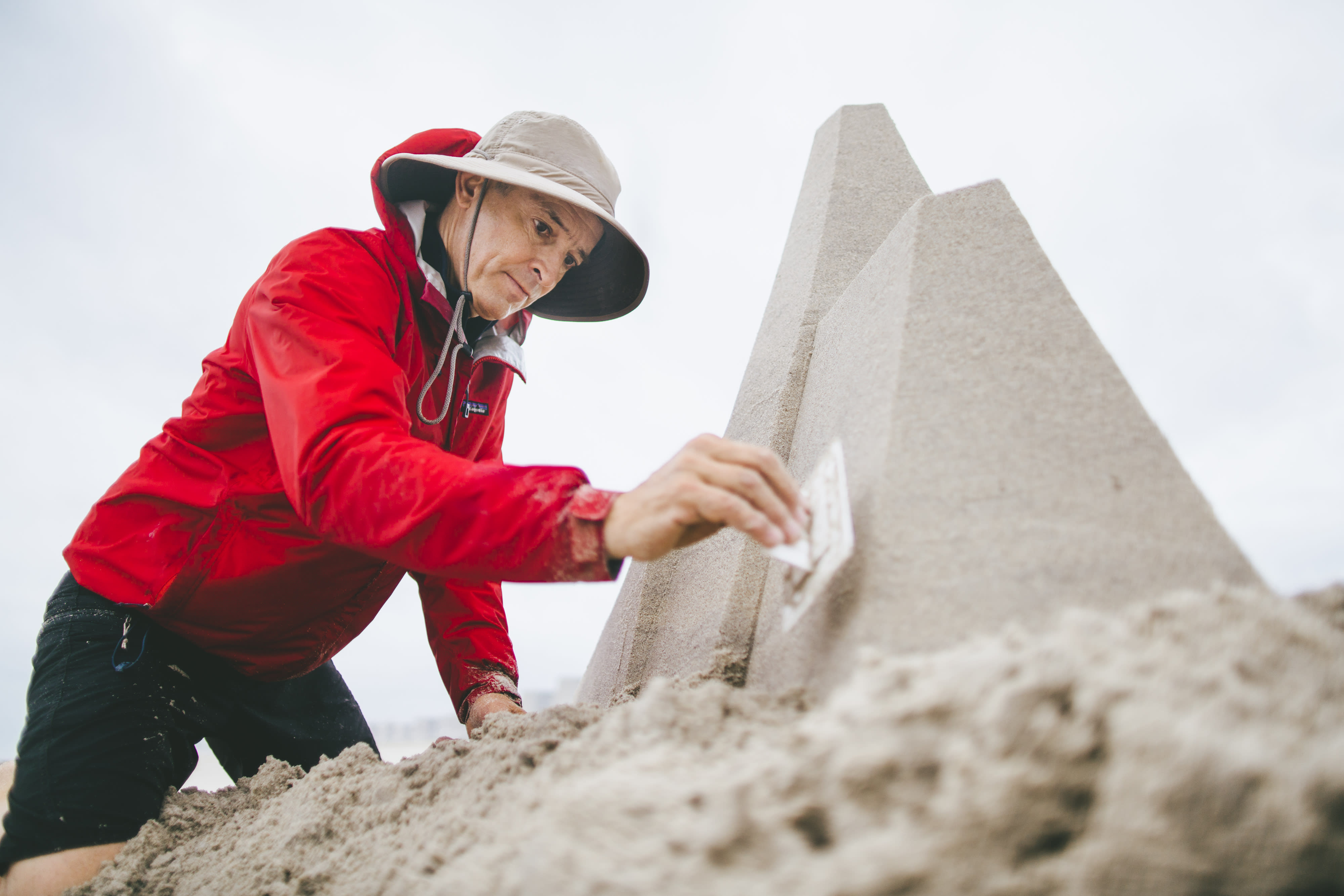 A man in a raincoat, hat, and shorts, kneels in the sand on a beach and works on a pyramidical sandcastle. He is tamping the sand down on one wall of the structure using a rectangular piece of plastic.