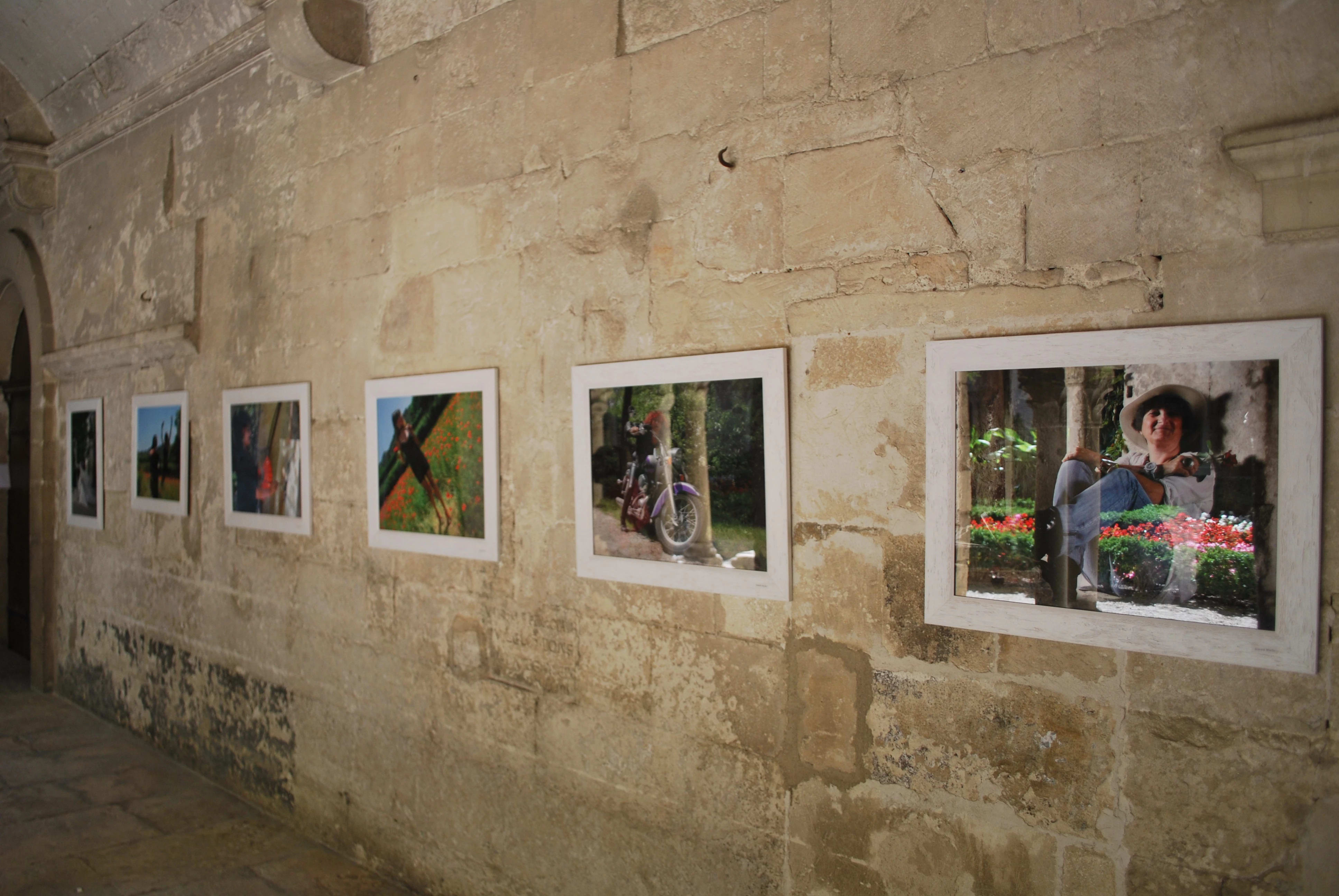 Many framed works hang in a row that runs the length of the stone hallway. The works are color photographs, and some of them feature people as their subjects.
