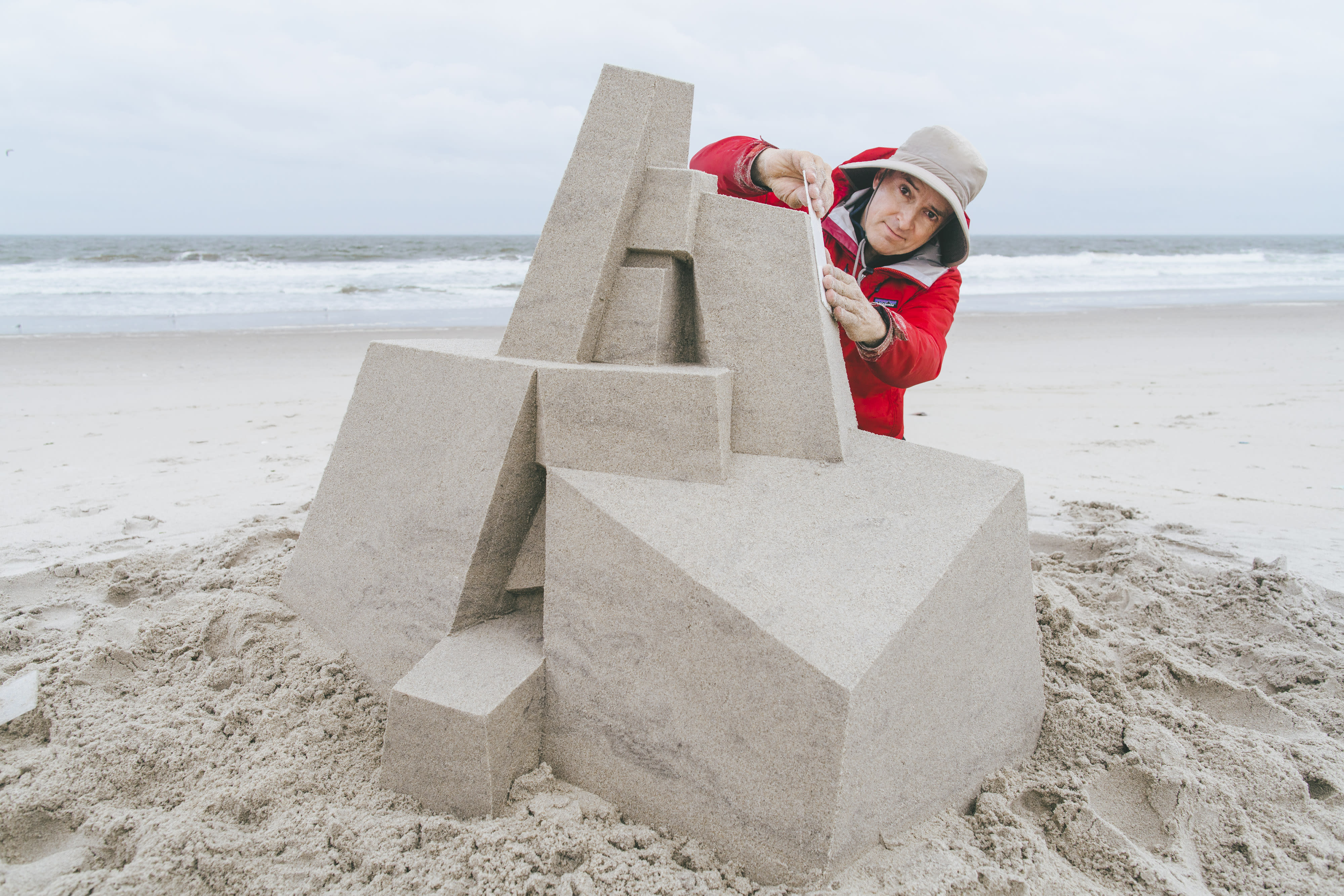 A man in a hat and raincoat on an empty beach puts the finishing touches on a sandcastle sculpture.