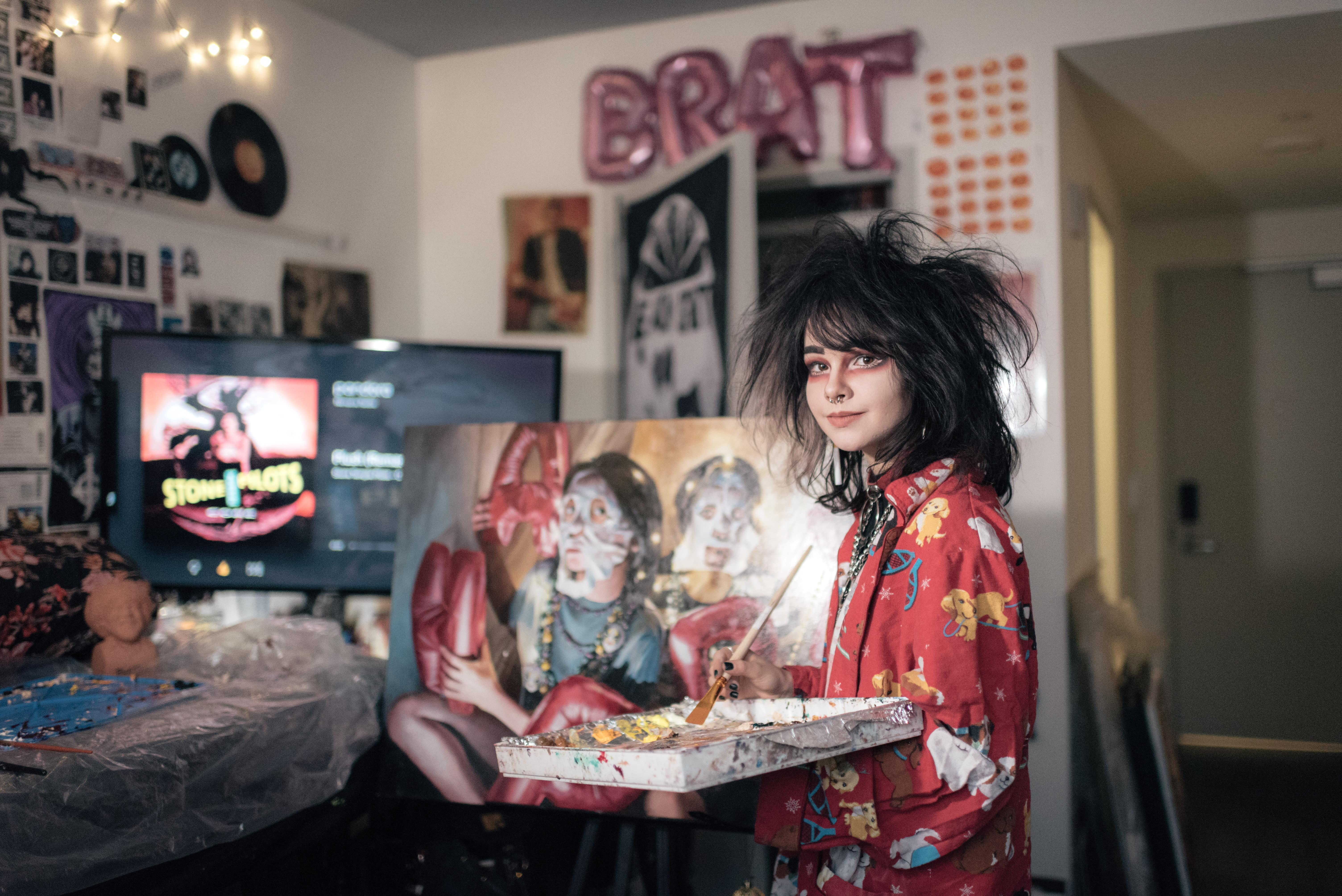 <p "="">A color photograph of a young woman holding a palette and paint brush and standing in front of a painting on an easel.
