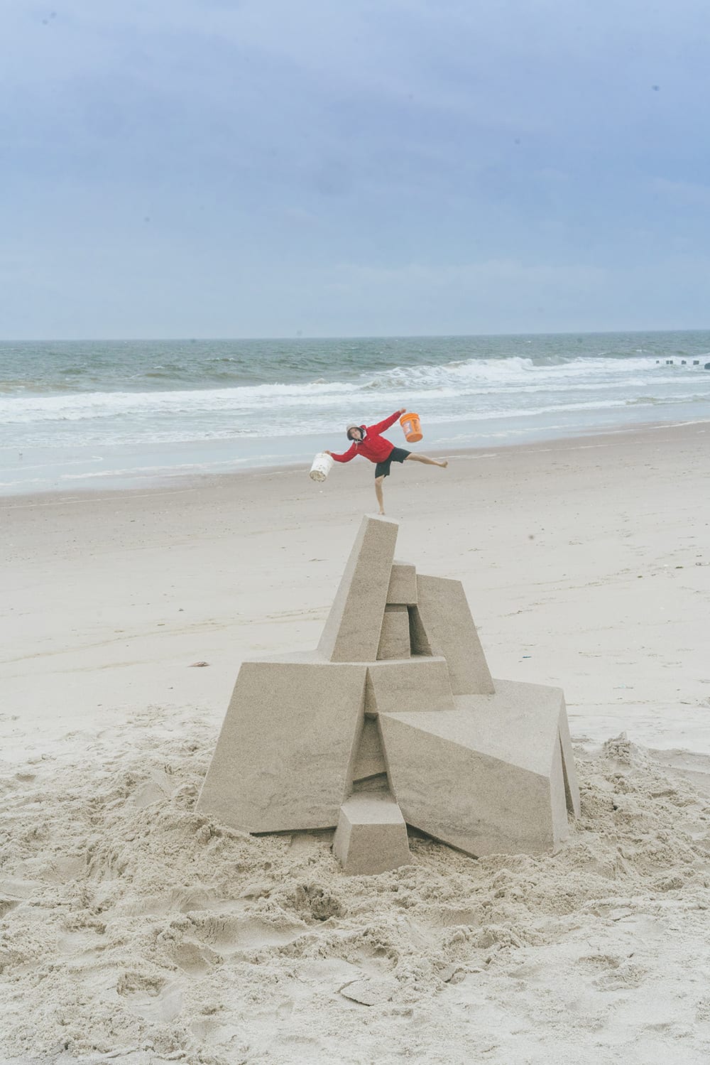 A man in a raincoat, hat, and shorts carrying a large bucket in each hand poses for a photo on an empty beach. In the foreground is a large sandcastle, and the man is posed so that he appears to be standing on the top of the sculpture.