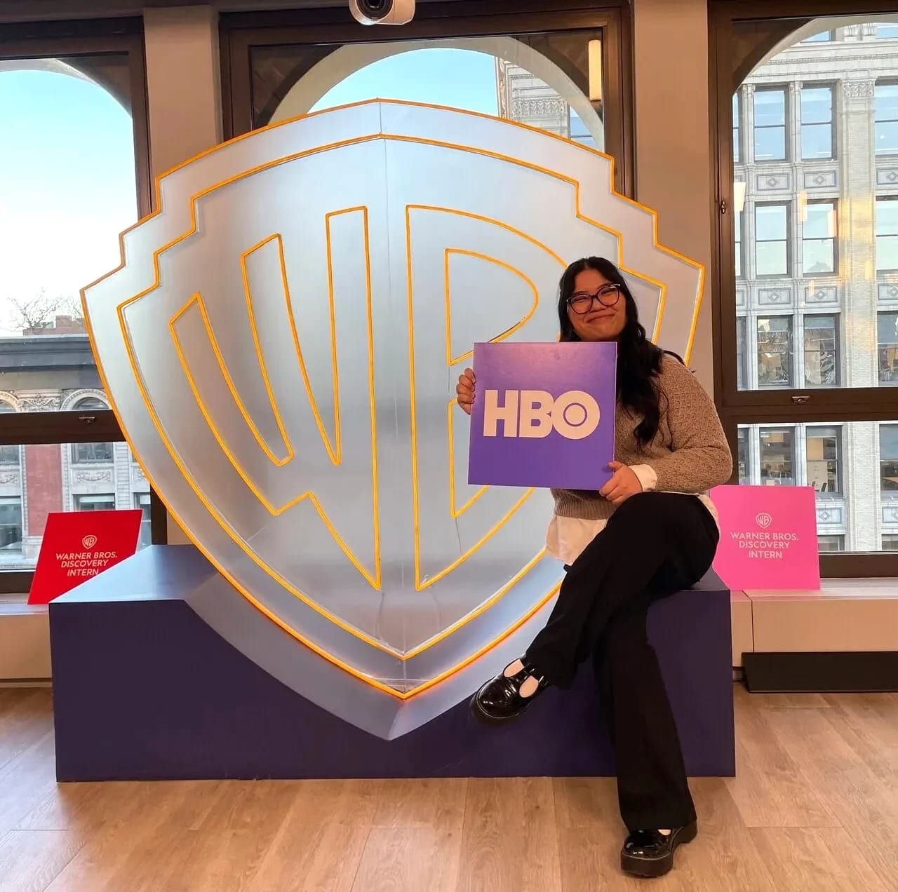 Student holding HBO logo sign in front of larger transparent Warner Brothers logo sign.