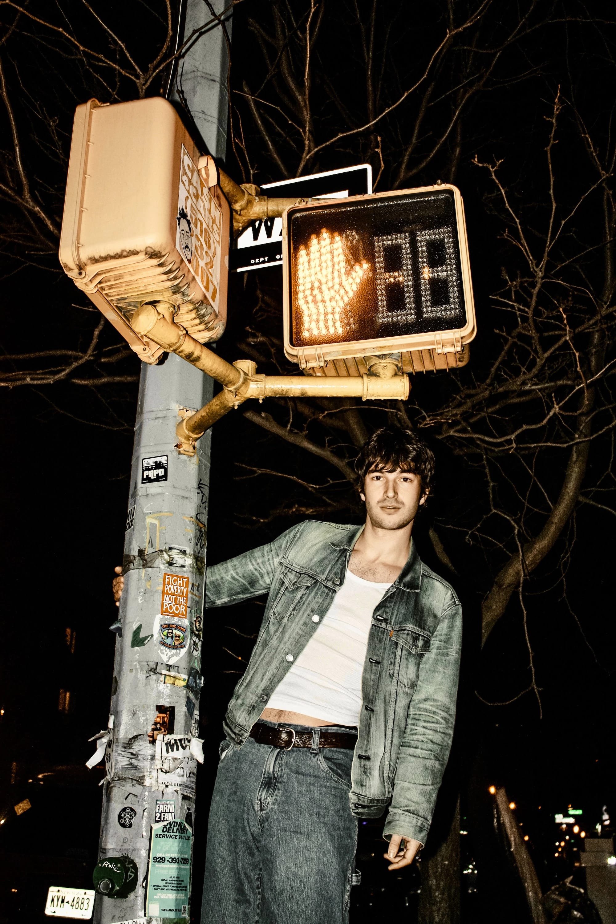 A portrait of man stood on top of, and hanging onto a streetlight at night.