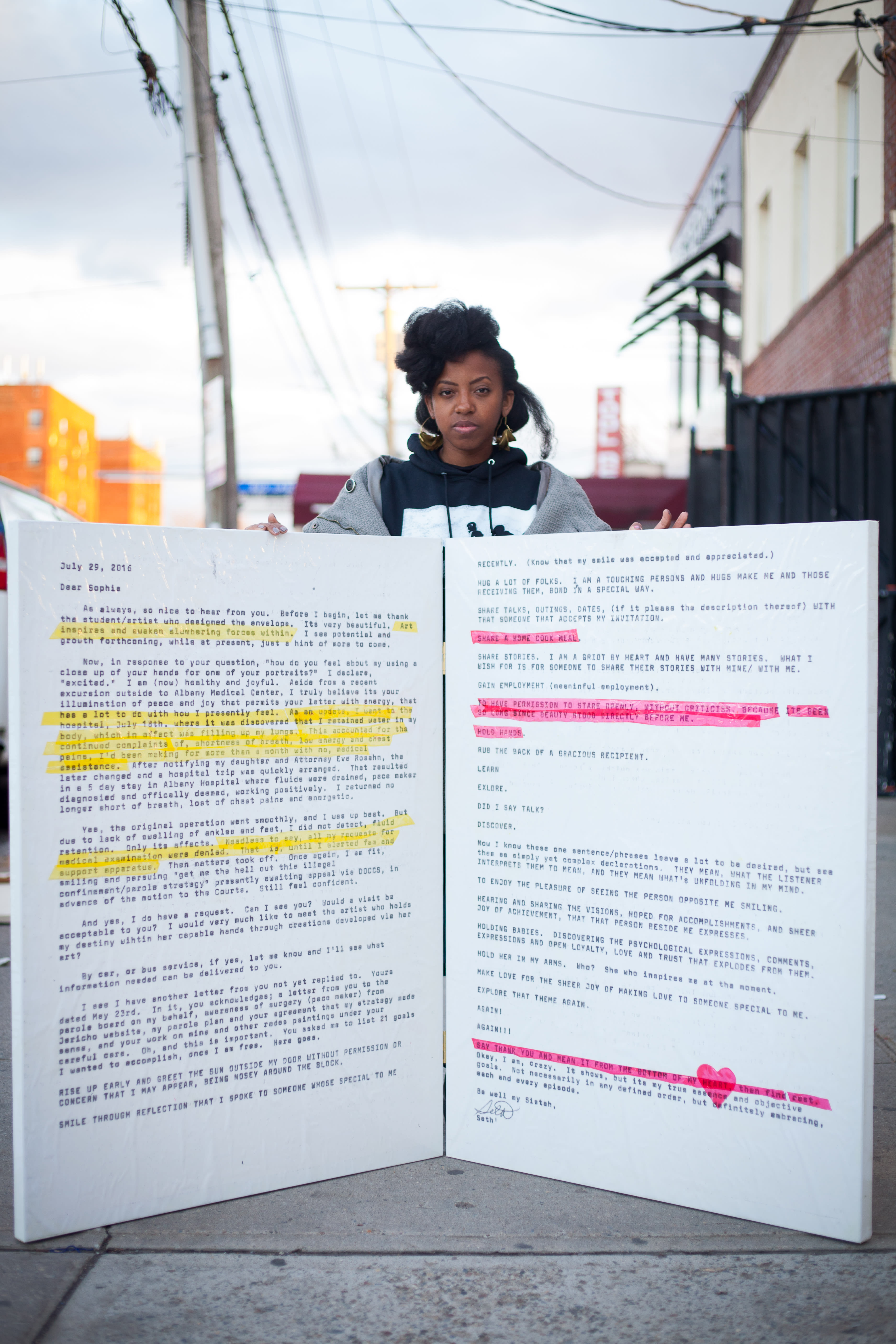 Artist Sophia Dawson standing on a city sidewalk holding a large poster with highlighted text.