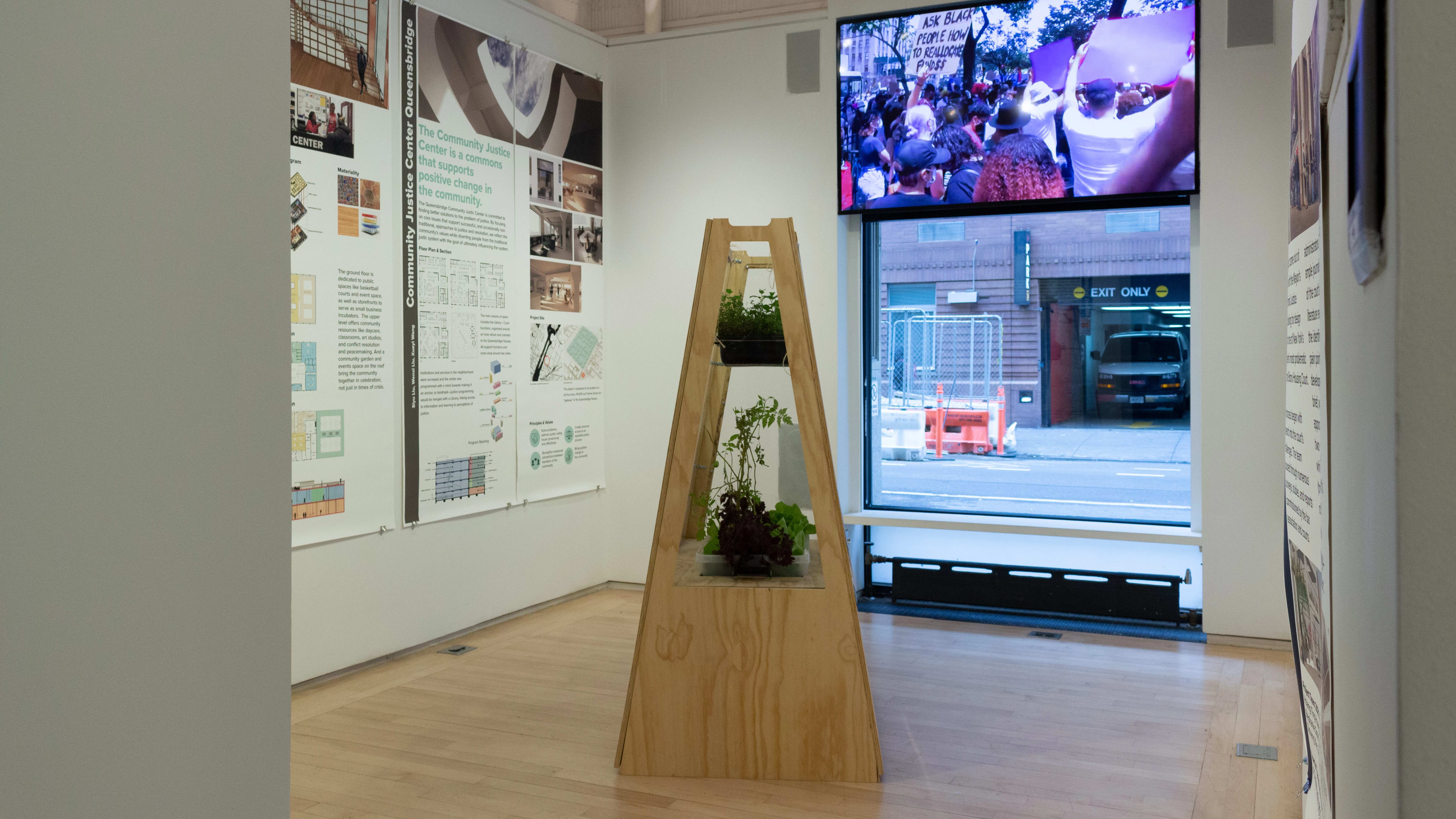 Exhibition view of An Infrastructure for Restorative Justice, featuring wooden sculpture with plants and large posters on the walls.