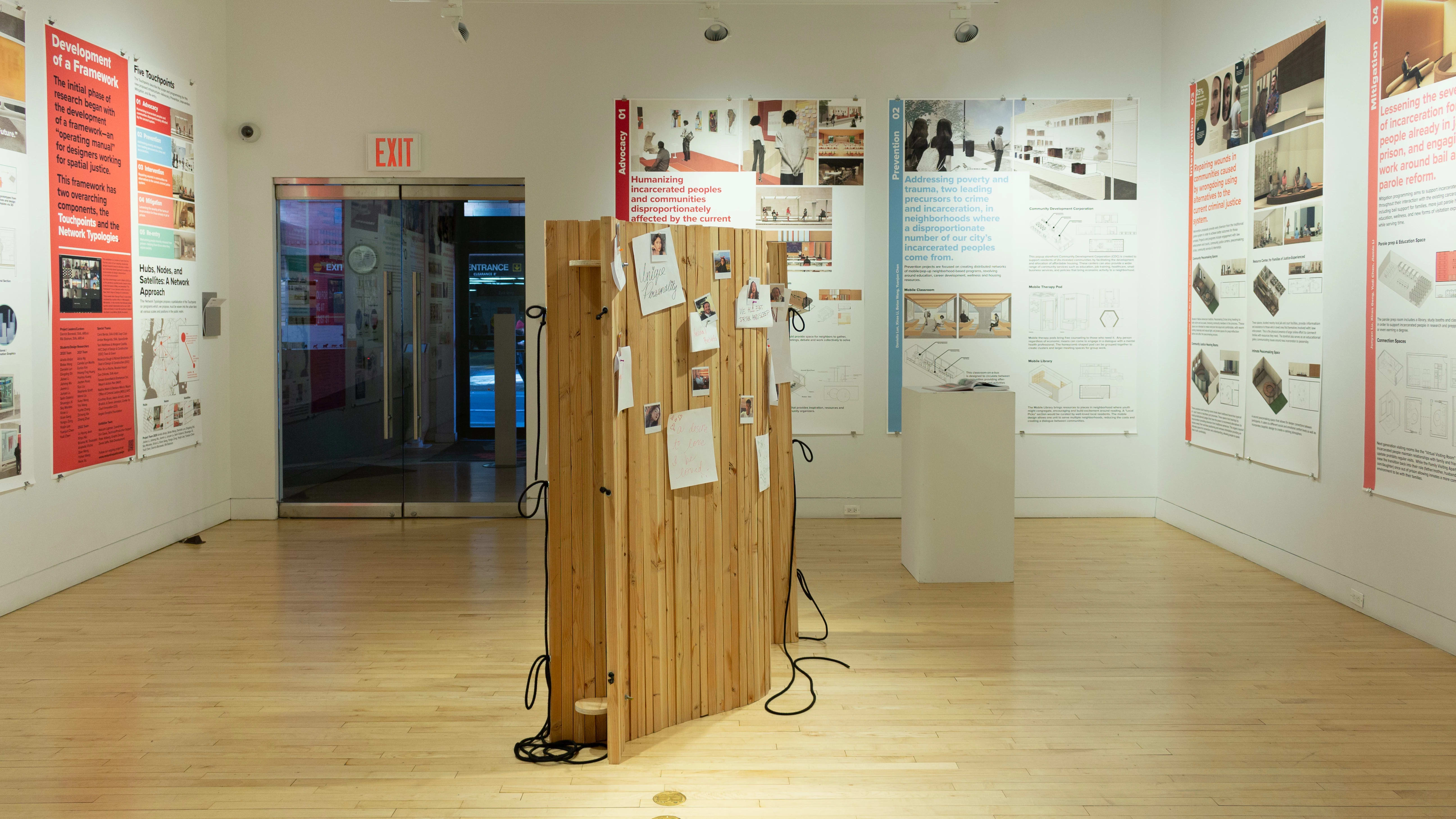Exhibition view of An Infrastructure for Restorative Justice, featuring wooden sculpture, pedestal, and large posters on the walls.