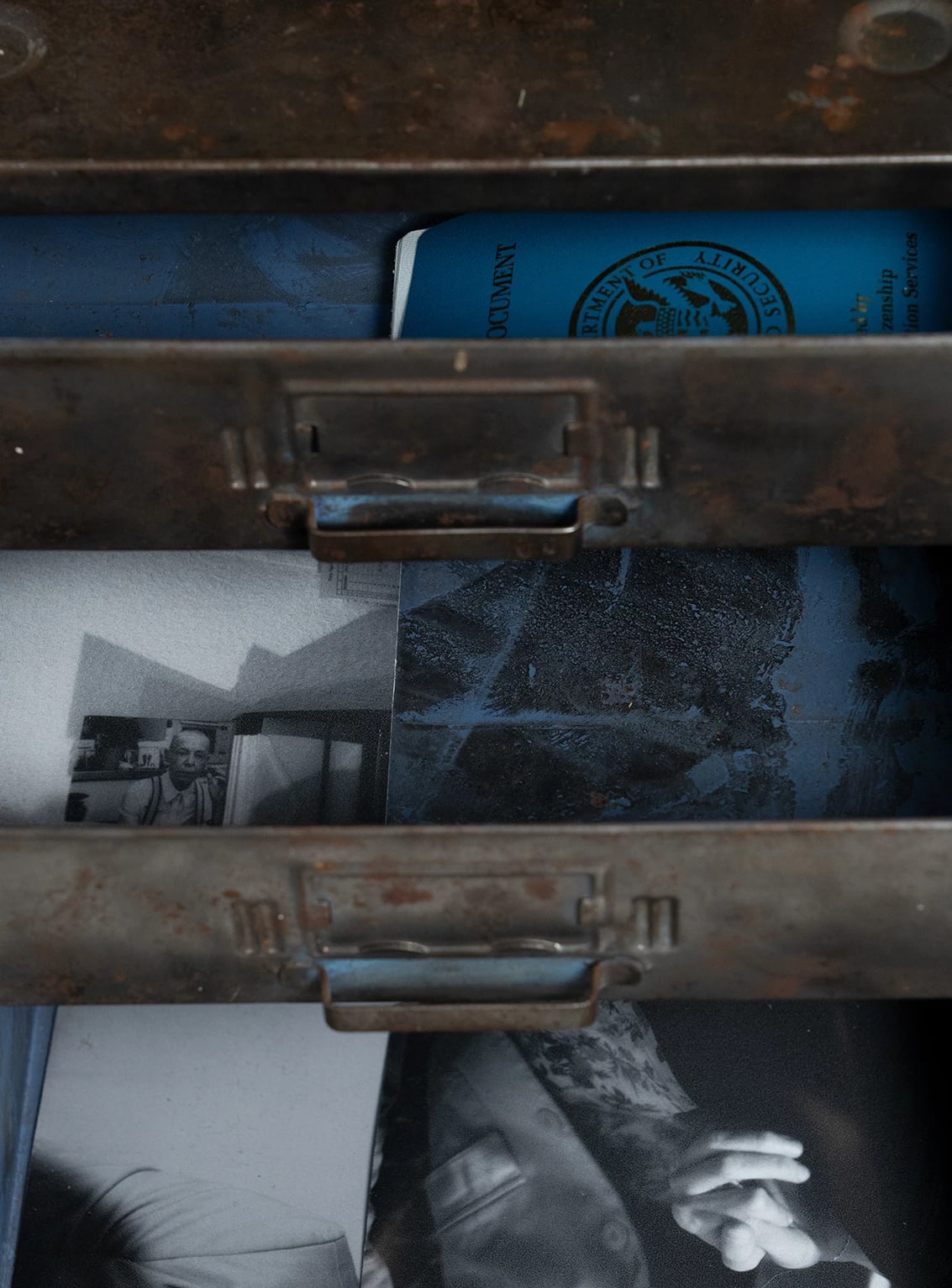 Color image of three stacked opened metal drawers containing black and white photographs and a blue passport.