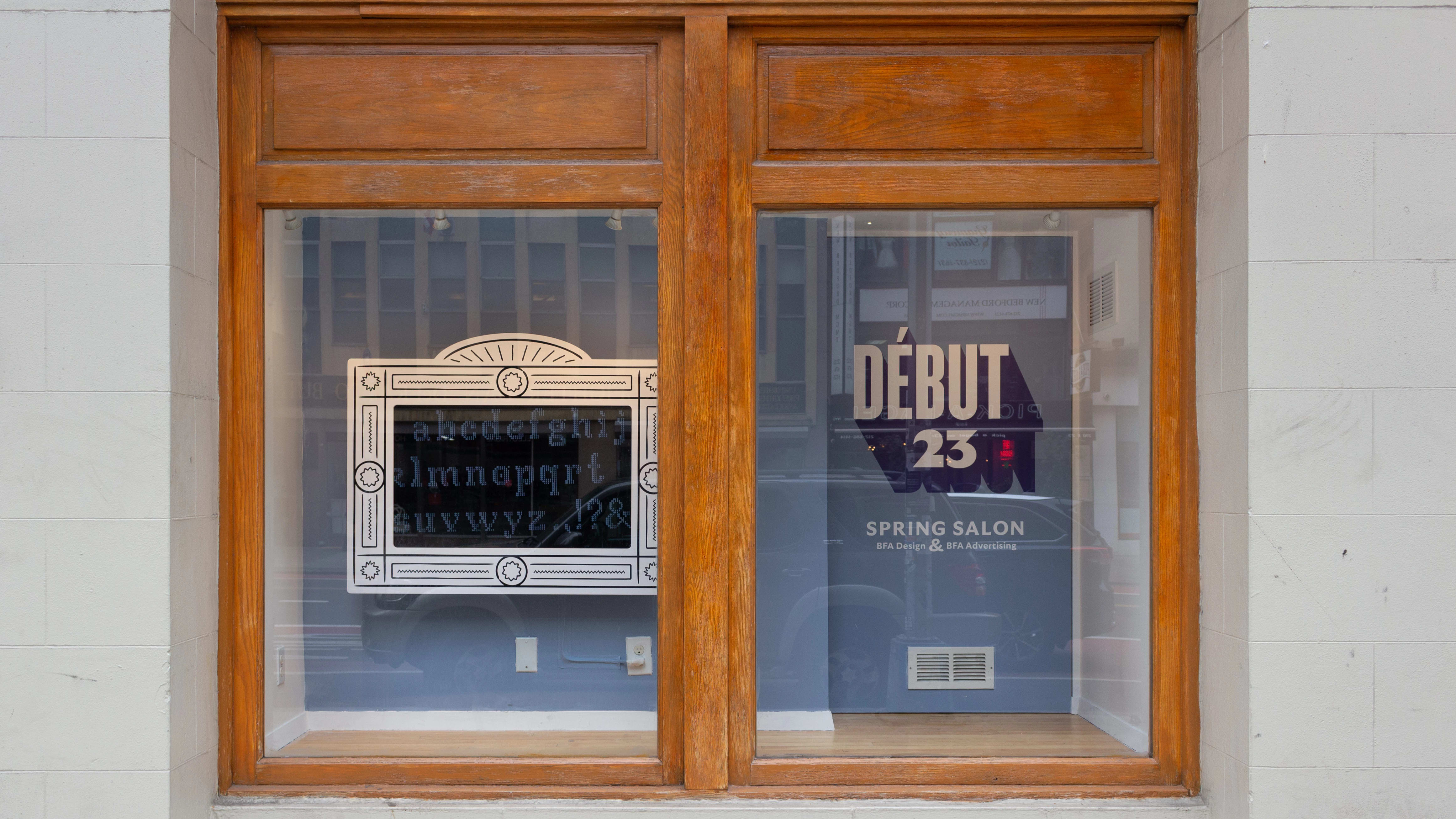 Installation image featuring a street view of a window with a framed monitor on the left and the show title, “Debut 23,” on the wall on the right in vinyl.