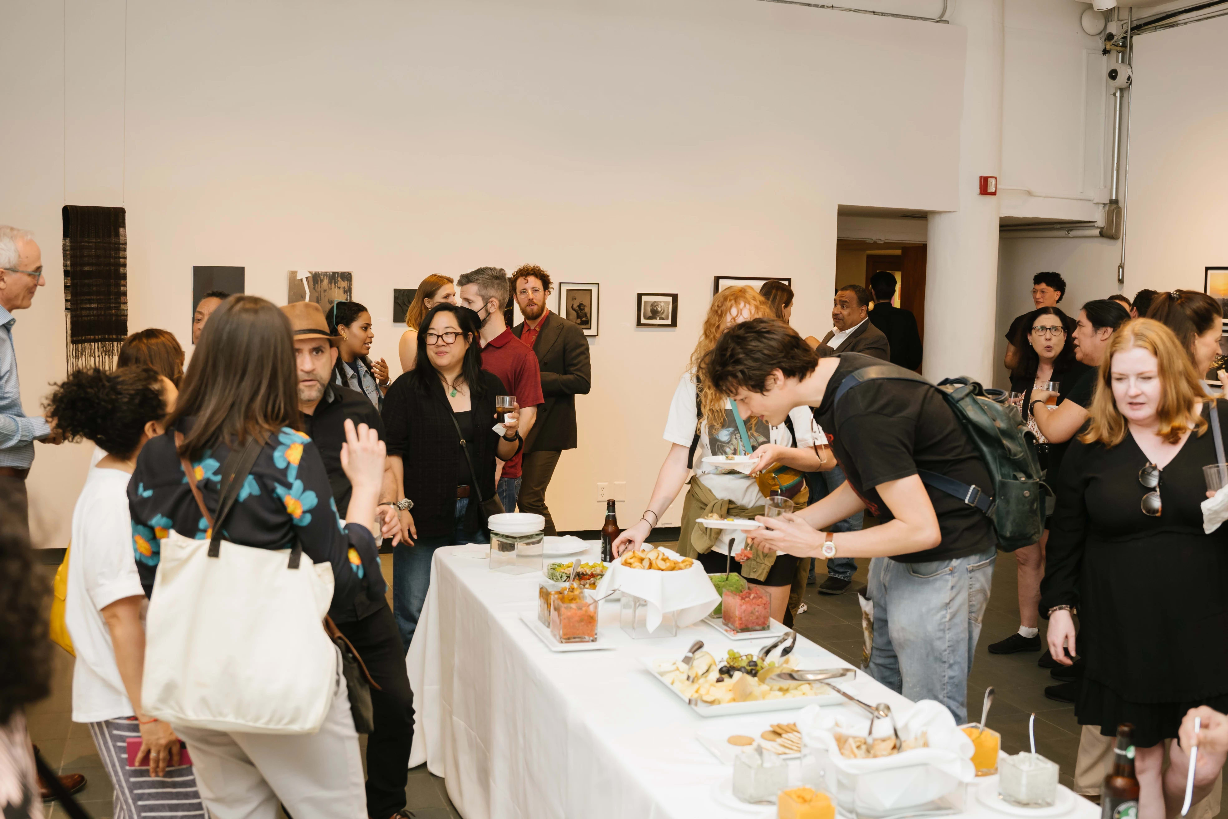 Reception photo showing many people at a gallery opening with a long food table in the middle.