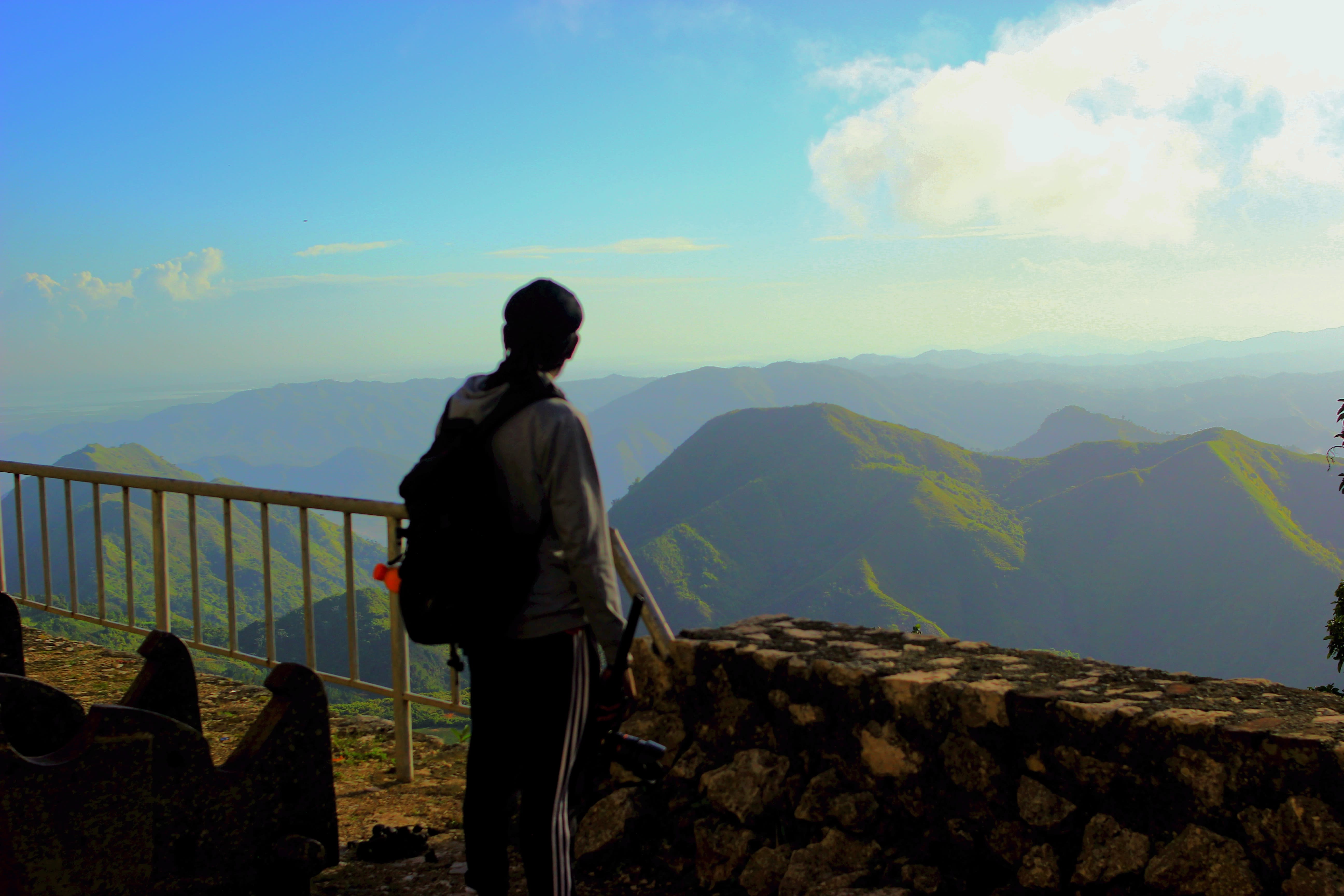 a man looks over a rocky half wall into a beautiful green hilly area and a bright blue sky.
