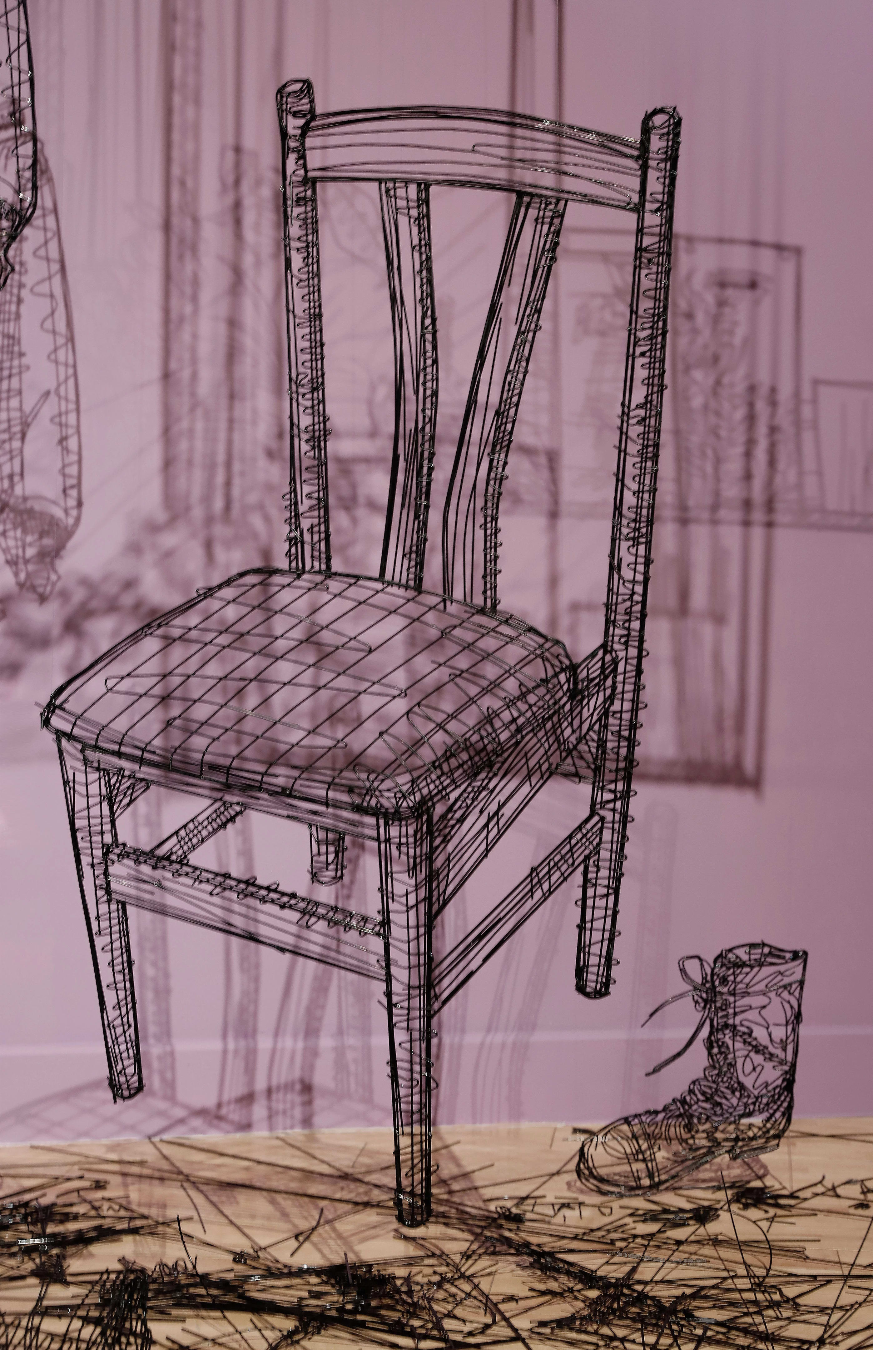Close up of a sculpture made out of black wire depicting a wooden chair with a small boot next to it.