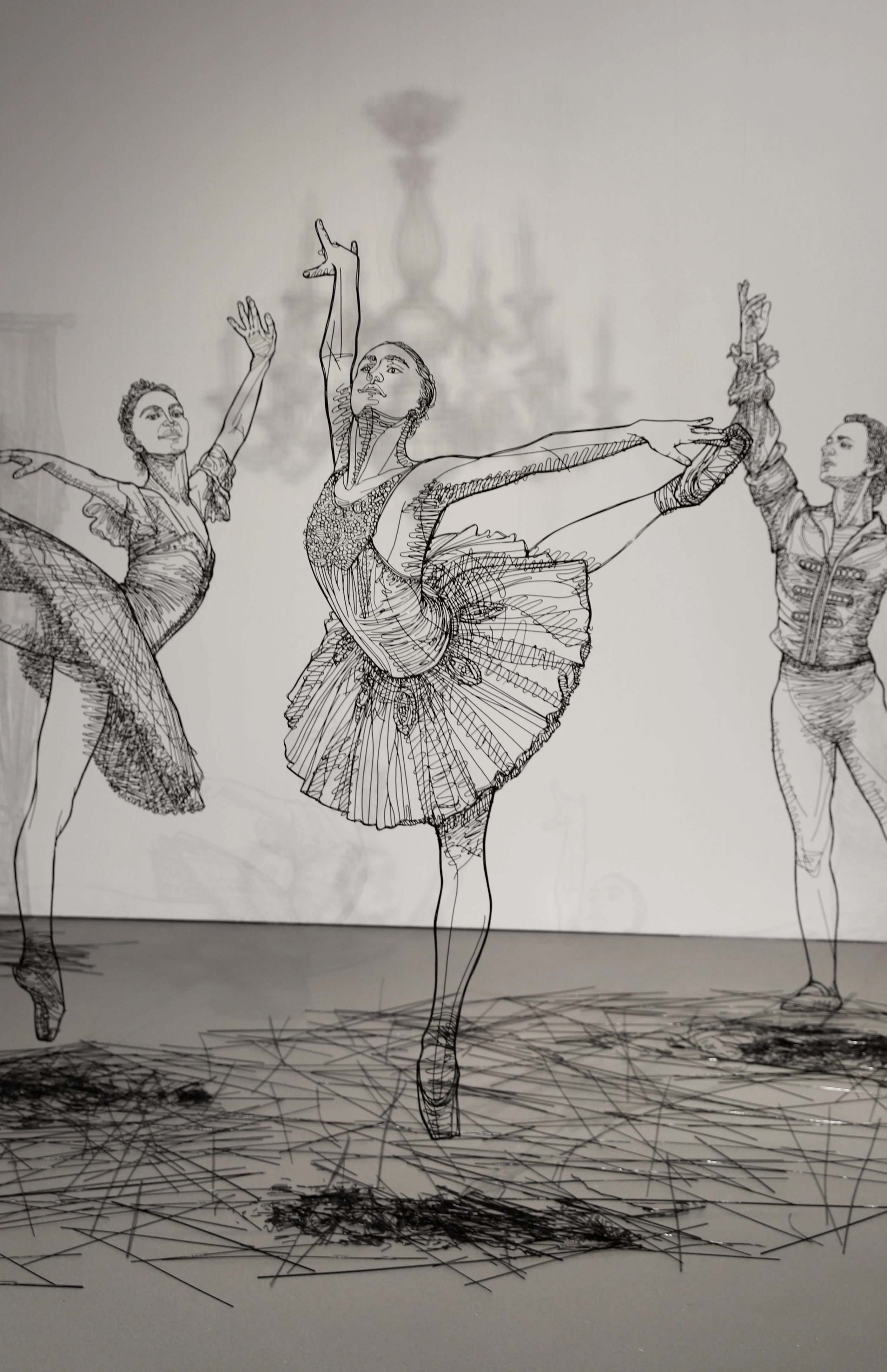 Close up of sculptures made in black wire of several large scale ballerinas dancing around a white room.