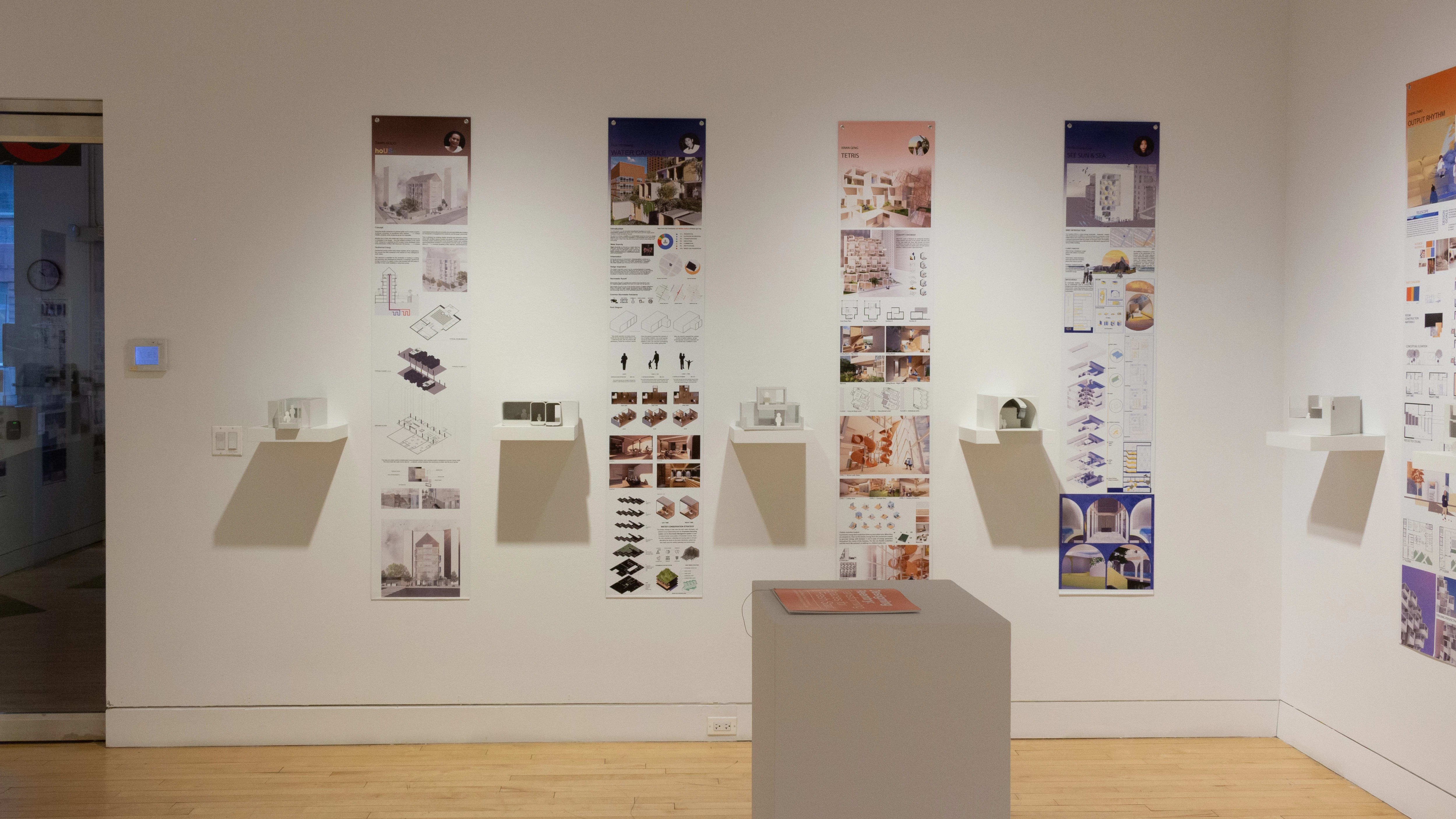 Installation image showing a straight view of two gallery walls with five building models on shelves in total, and information banners for each to the right. There is a pedestal on the floor with a book on top.