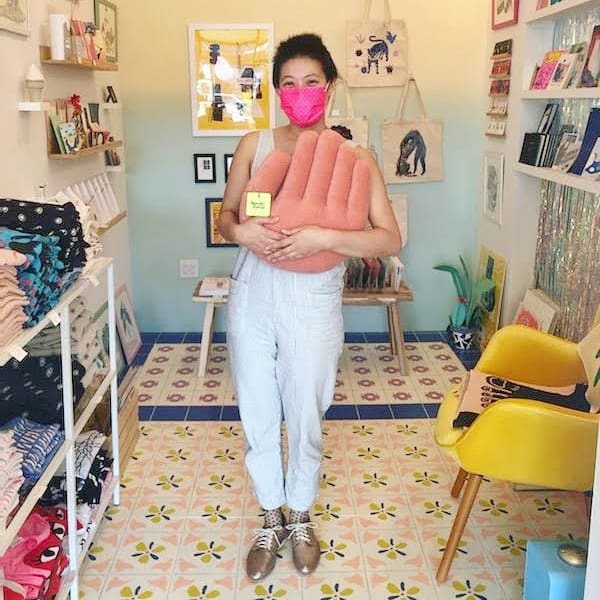 A photograph of a person wearing a face mask, holding a hand-shaped throw pillow and standing in a shop with a colorfully tiled floor.