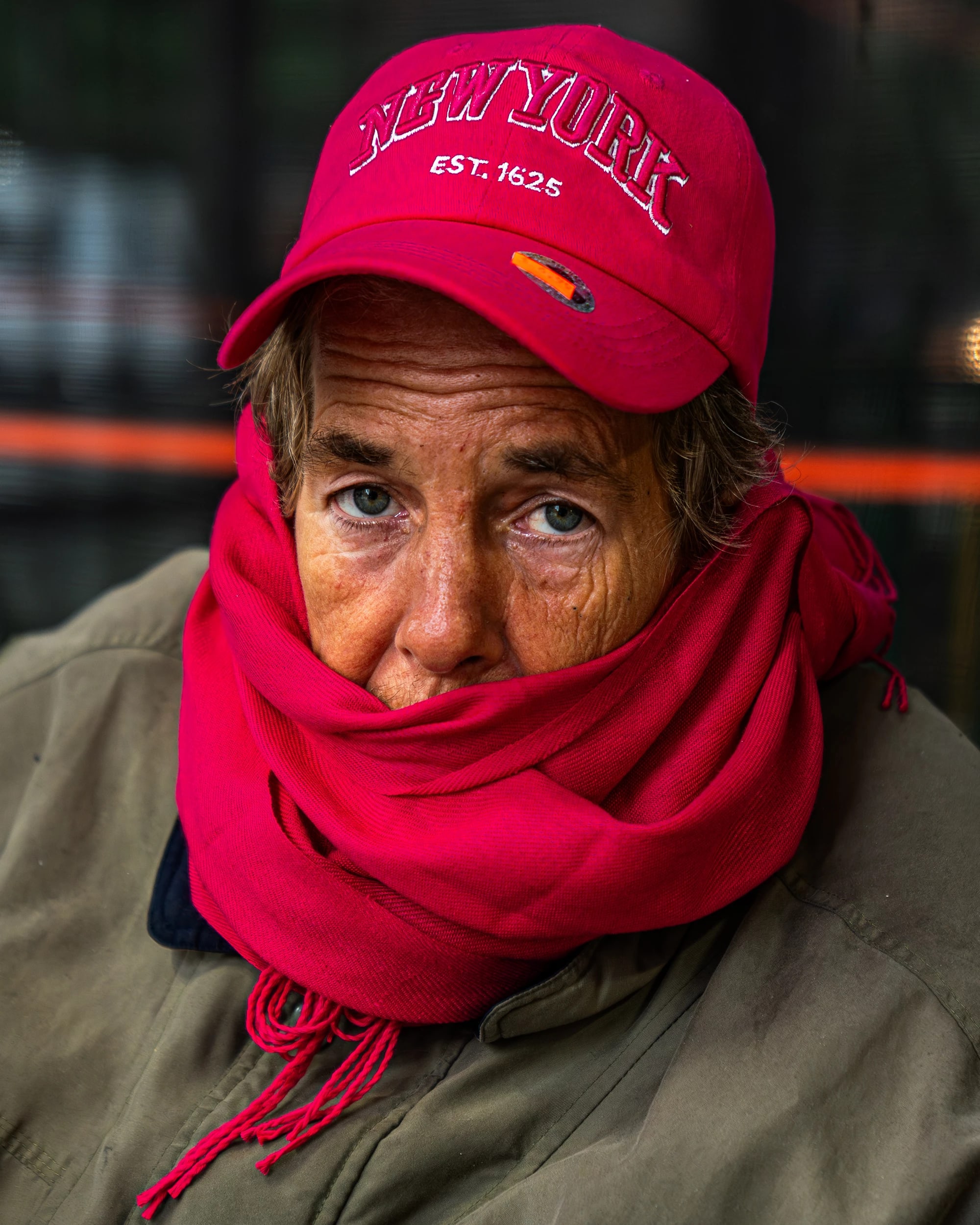 A portrait of an elder person with blue eyes and wrinkles on their face. They wear a red hat, red scarf and green coat.