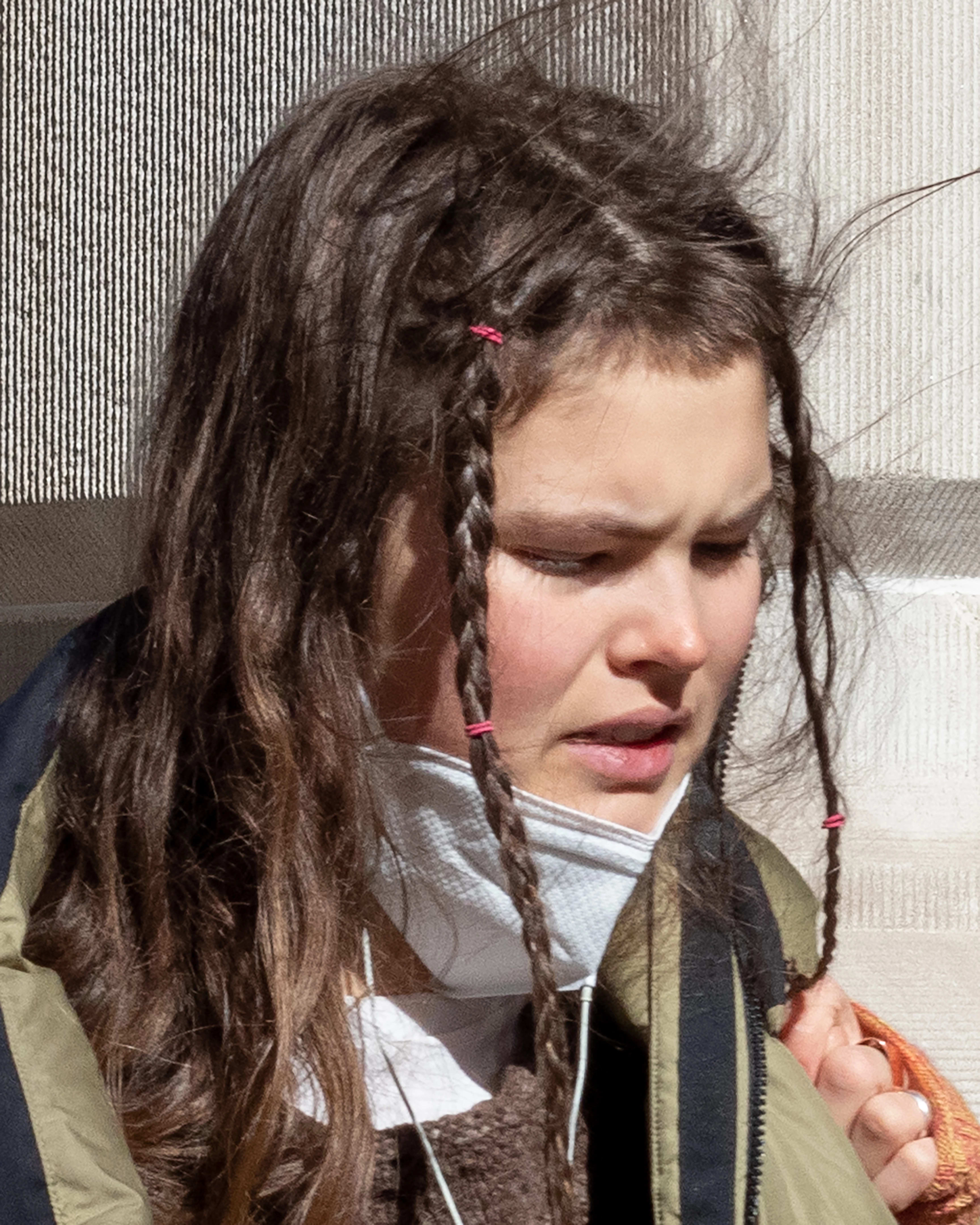 45-degree angle portrait in color of a woman looking downward with a white KN-95 mask on her chin and braids framing her face, close-cropped from the chest up, with a light tan stone facade in the background.