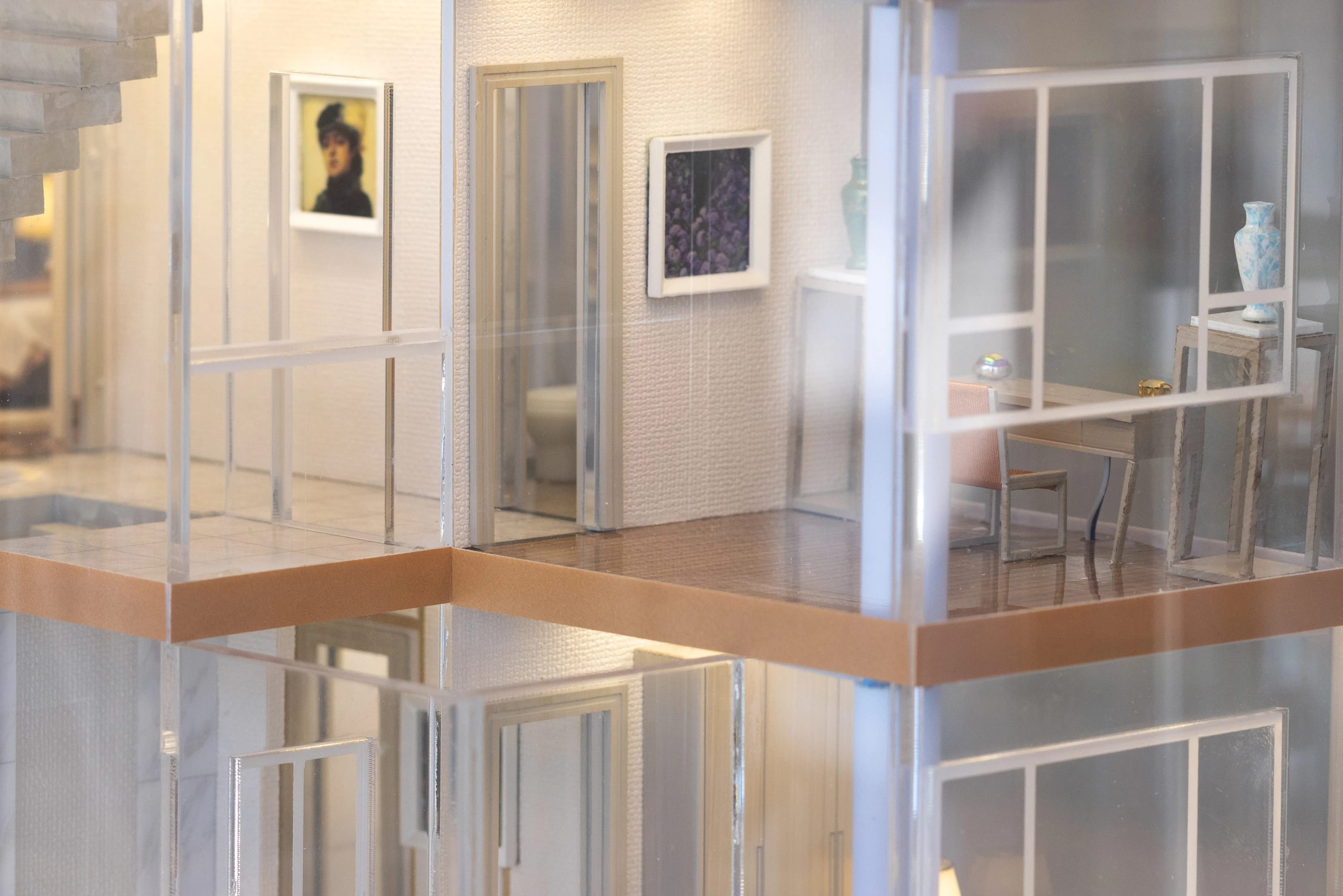 A color photograph  of a miniature of an apartment interior, decorated with two oil painting on the wall, the bathroom door is slightly open. On the right corner there is a desk, a chair and two vases.