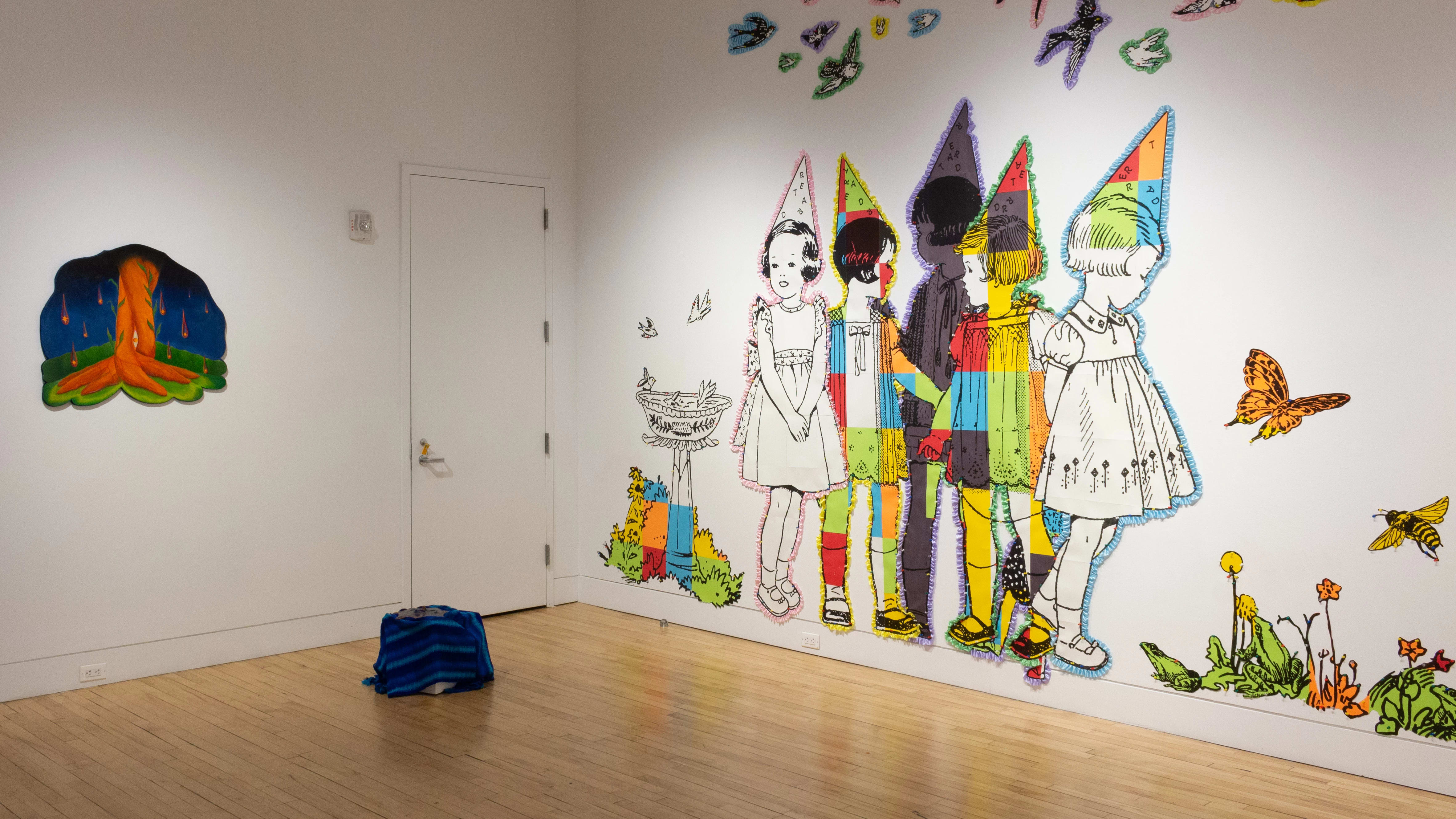 Next to the paper children on the back wall, a small box covered in a blue textile sits on the floor. On the wall to its left is an orange, blue, and green painting on a cut wood panel.