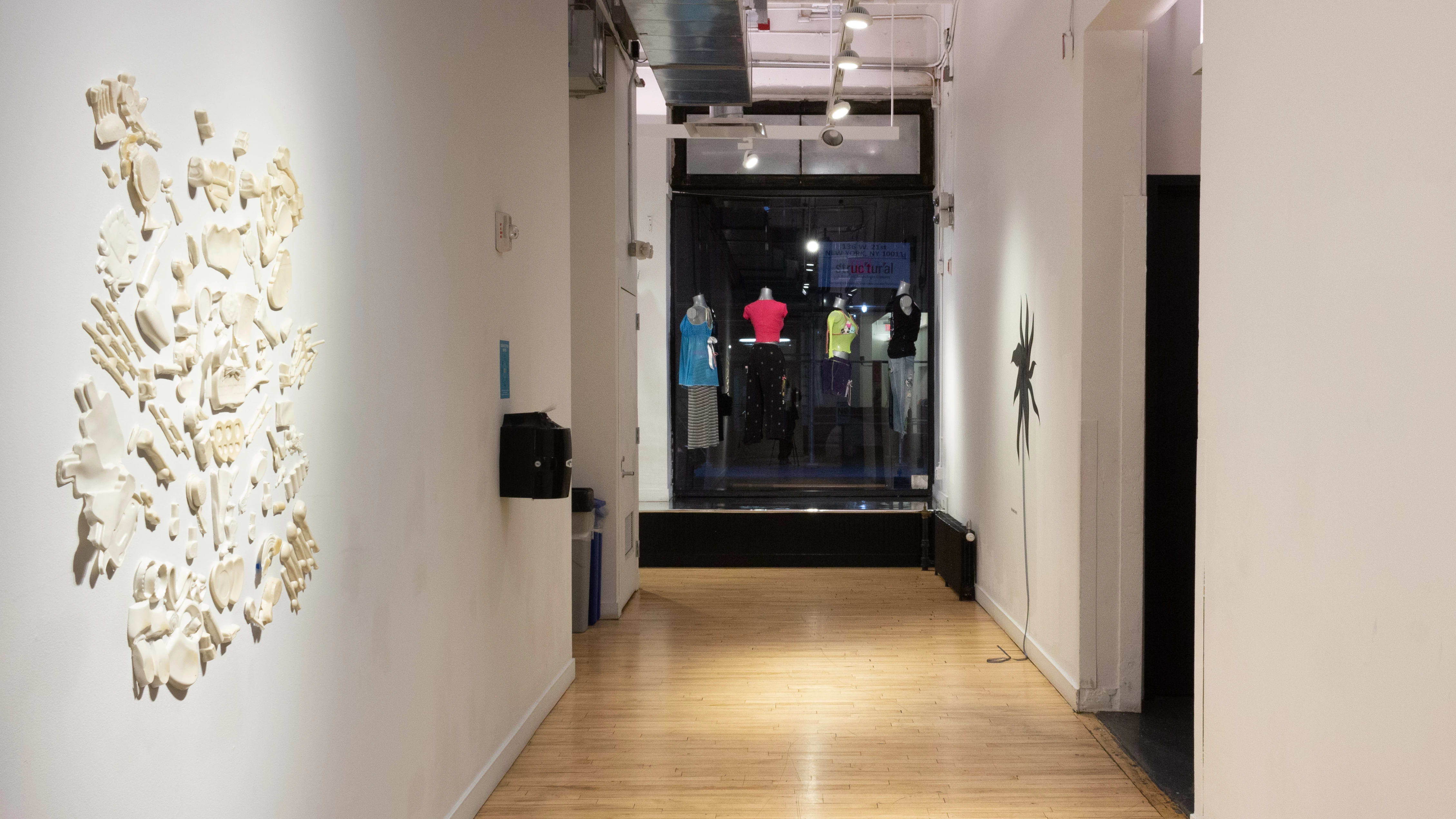 In front of a window, mannequins hang from the ceiling dressed in different clothing. On the left wall, white and cream colored objects are arranged in a cluster.