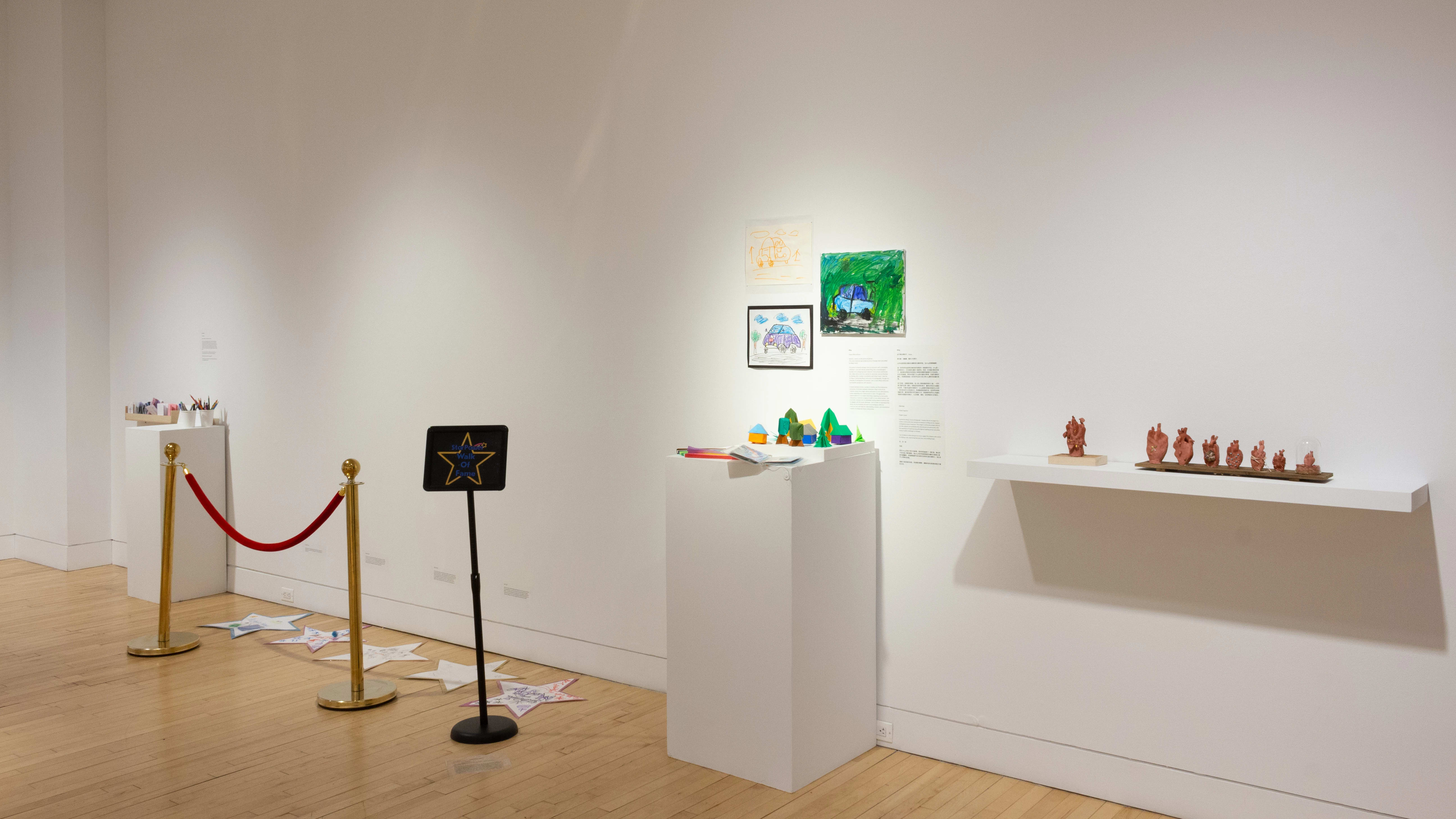 Against one wall of the SVA Flatiron Gallery, a shelf holds several small sculptures of an anatomical heart. To the left of the shelf, colorful paper works sit on and above a pedestal. Further left, a stanchion with a red velvet rope protects a row of paper stars that lay on the ground.