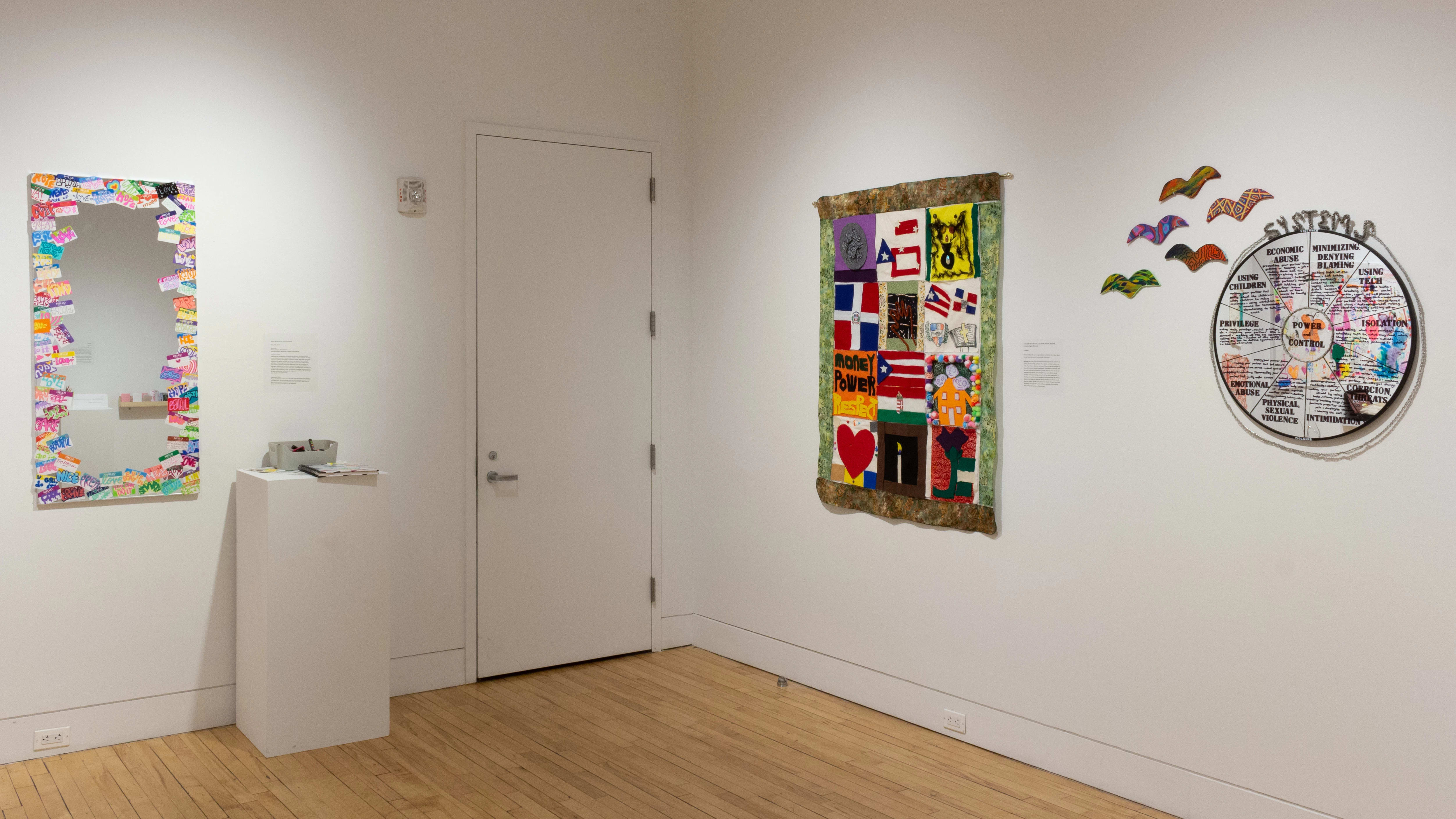 A circular mirror on the wall is divided into sections titled with various systems of power and control. Left of the mirror, a quilt contains imagery like a heart, various countries’ flags, and the words “MONEY POWER RESPECT.”