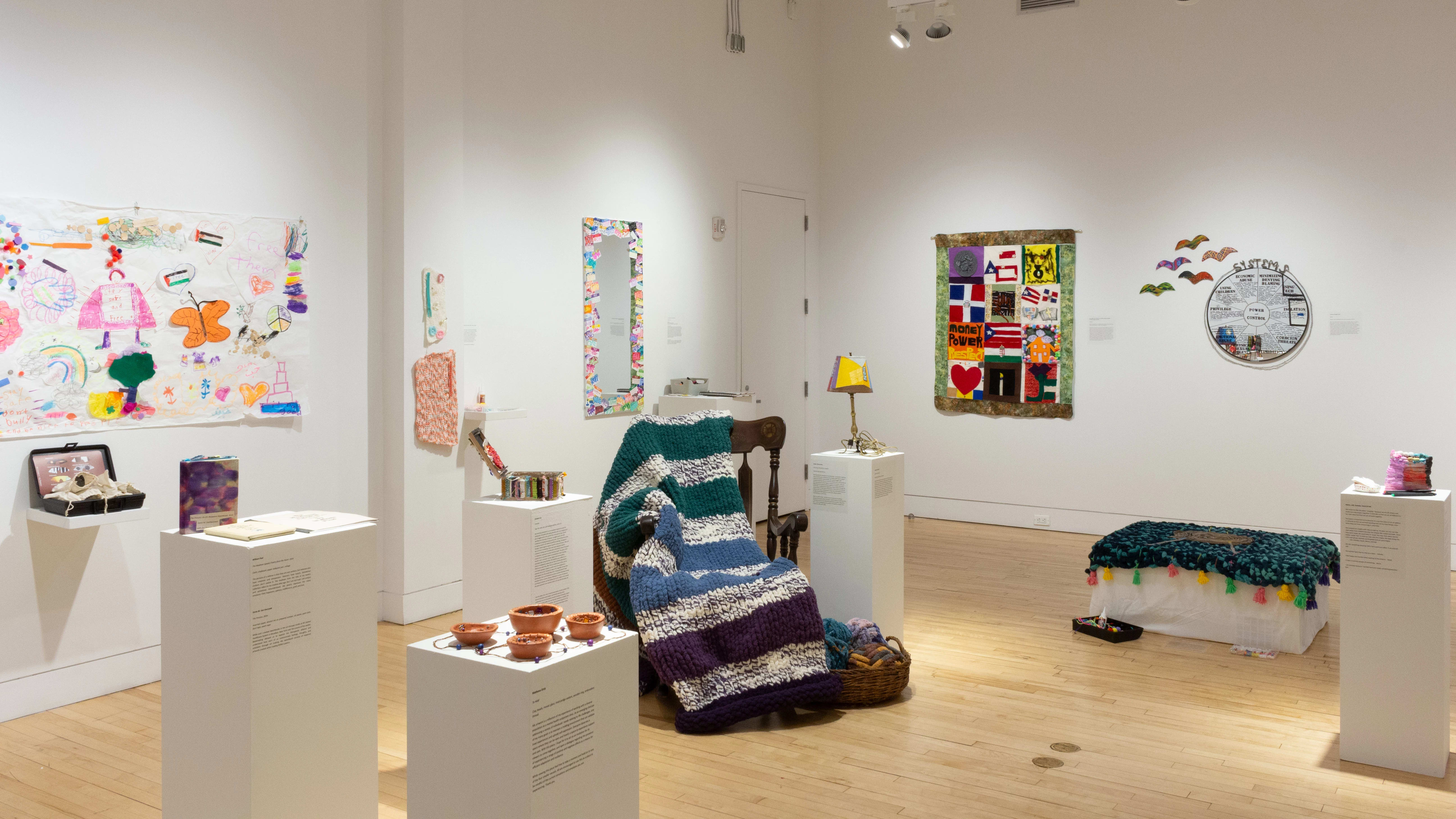 A wider view of the gallery shows a chair with a knit blanket across it, surrounded by pedestals that hold small sculptures. One wall holds colorful drawings and a mirror with a vibrant frame.