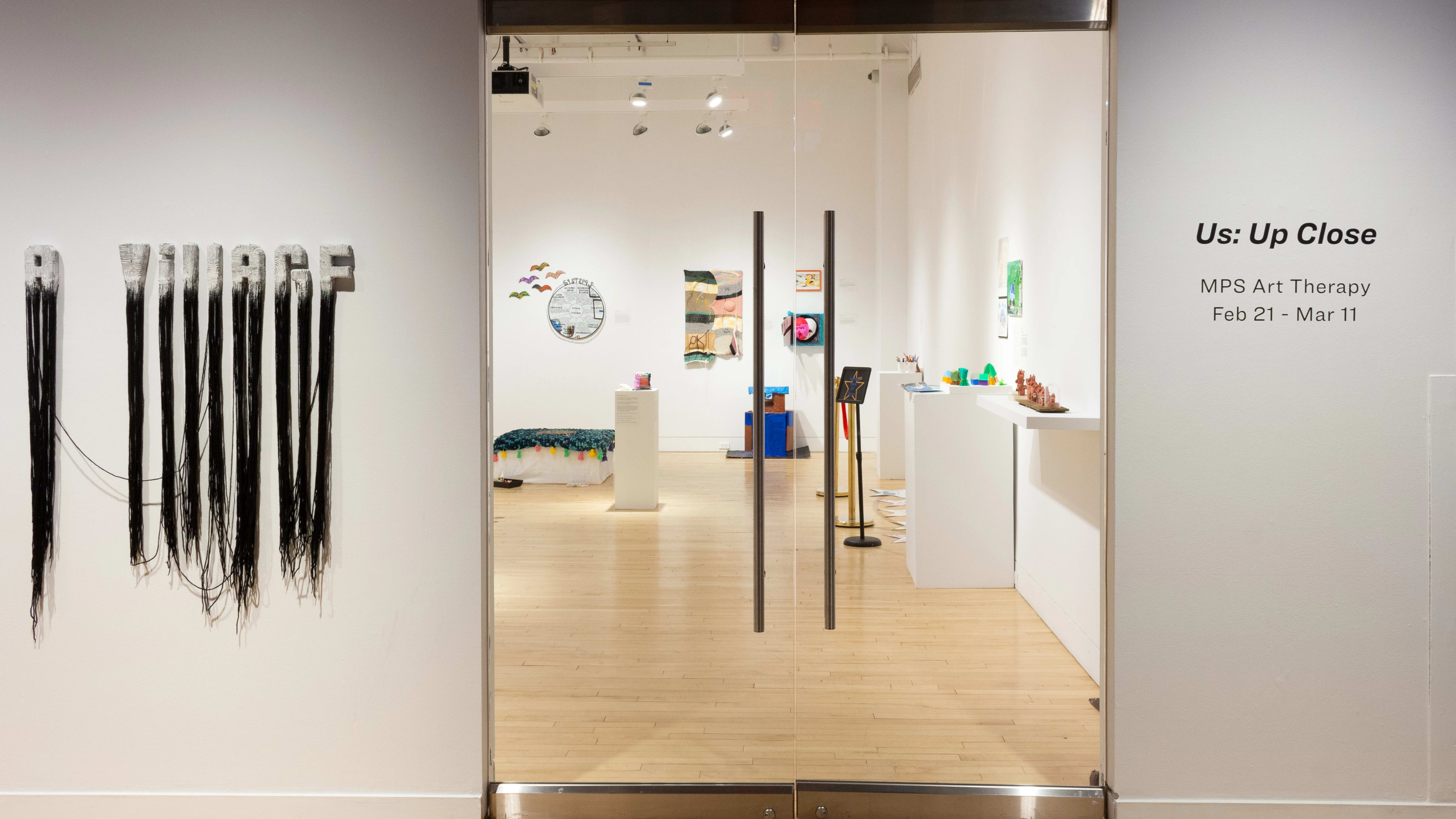 A sculptural piece that reads “A VILLAGE” hangs to the left of the door into the gallery. To the right of the door, wall vinyl shows the exhibition title “Us: Up Close.”