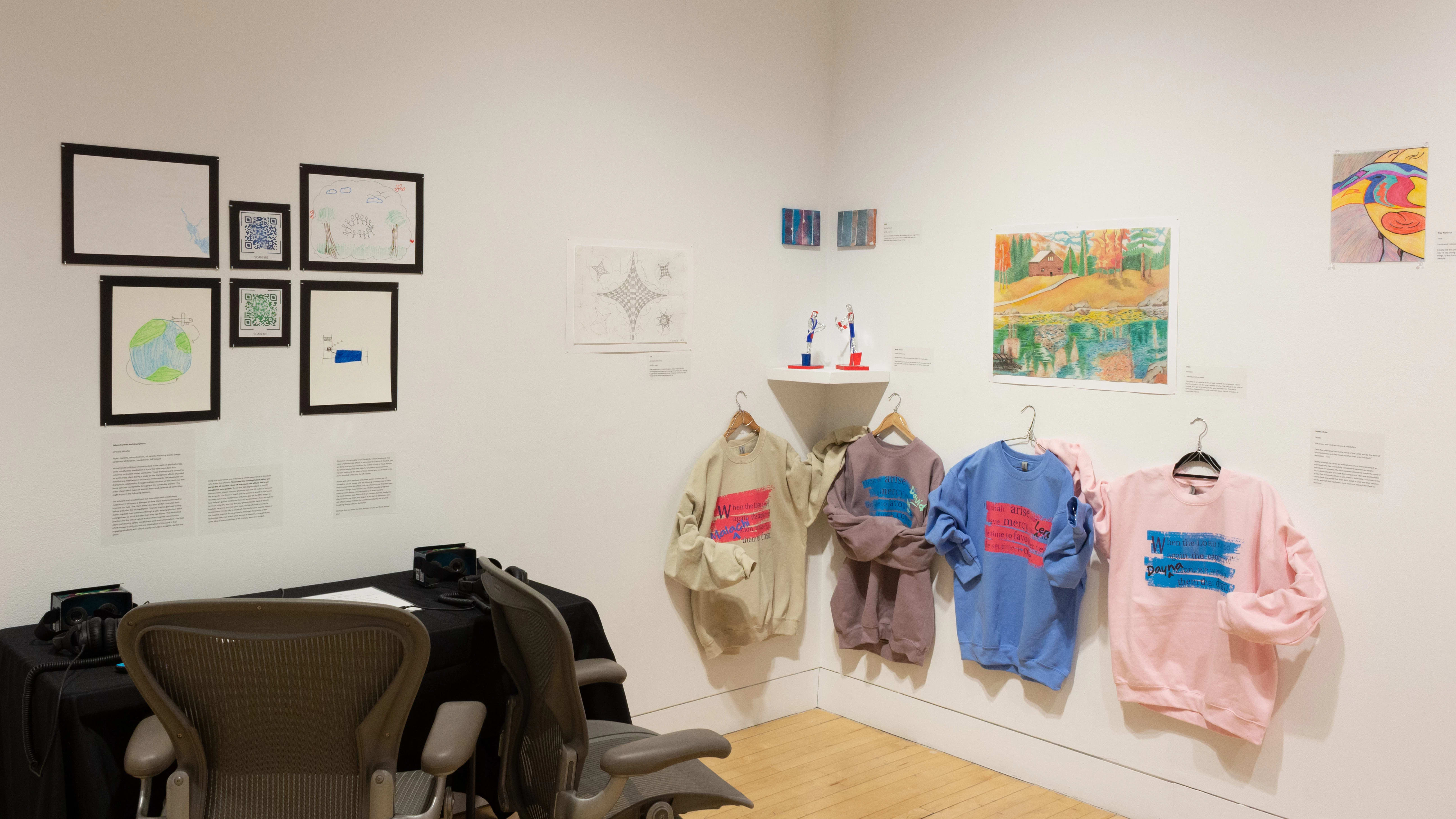 In one corner of the gallery, there are two office chairs facing a table and several framed works hanging above it. To the right, a row of sweatshirts are printed with pink and blue text. Around them hang various drawings and small sculptures.