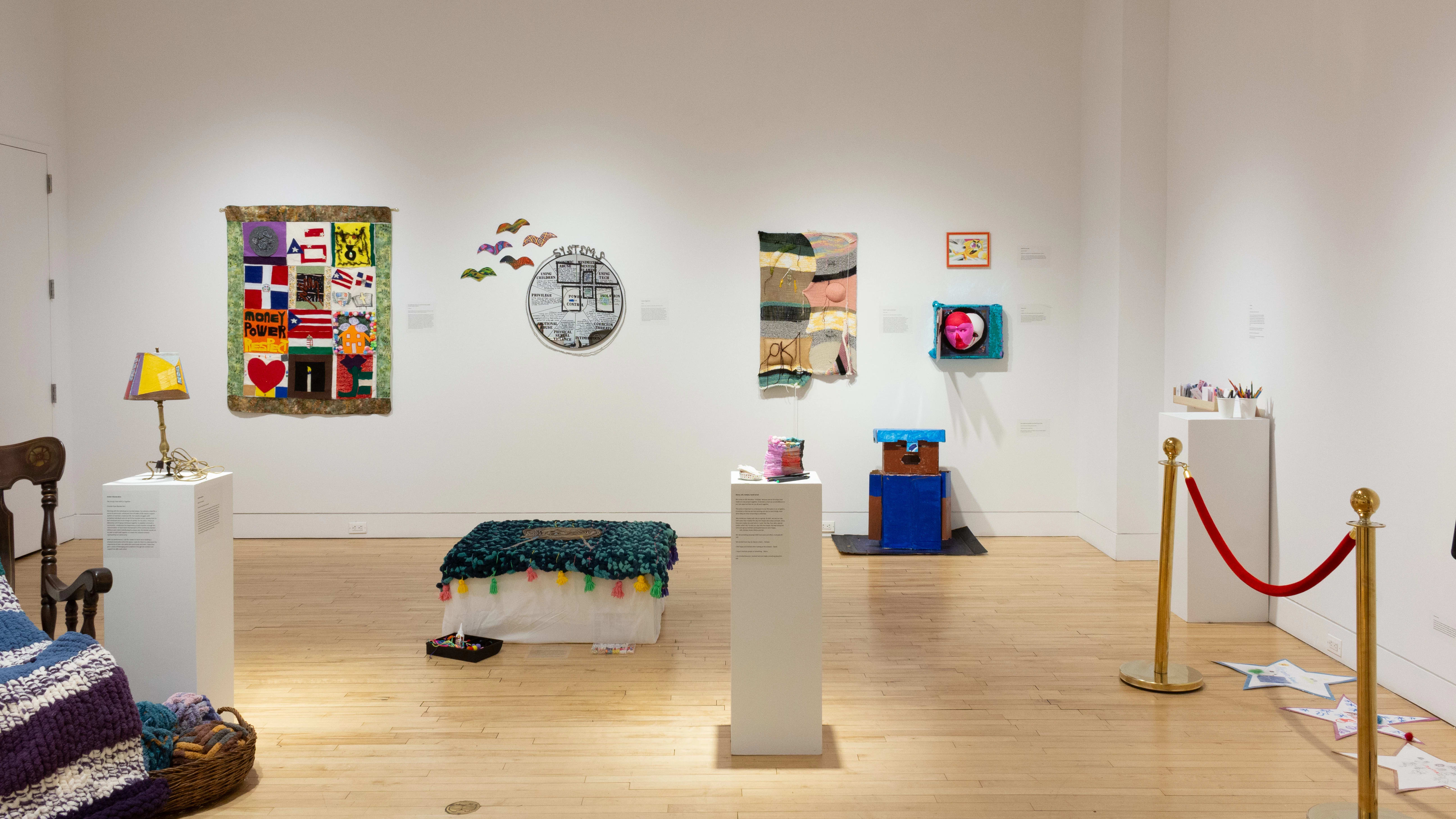 A view of the gallery shows the rocking chair, quilts, ottoman, lamp, paper stars, and stanchion, in addition to other works of various mediums.