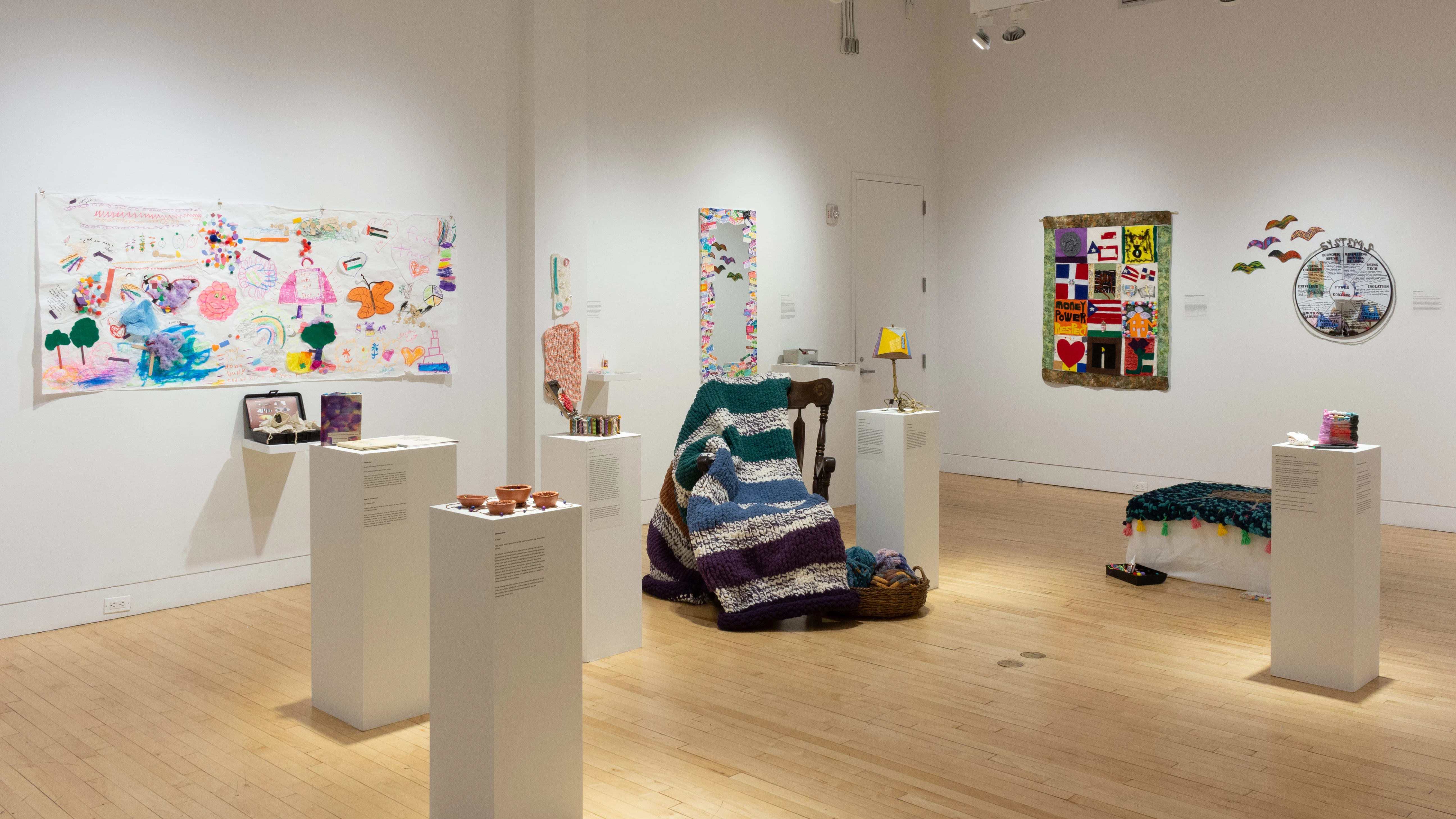 Pedestals holding small sculptures surround a rocking chair and blanket, which sit in the center of the room. Colorful drawings and textile works hang on the walls behind them.