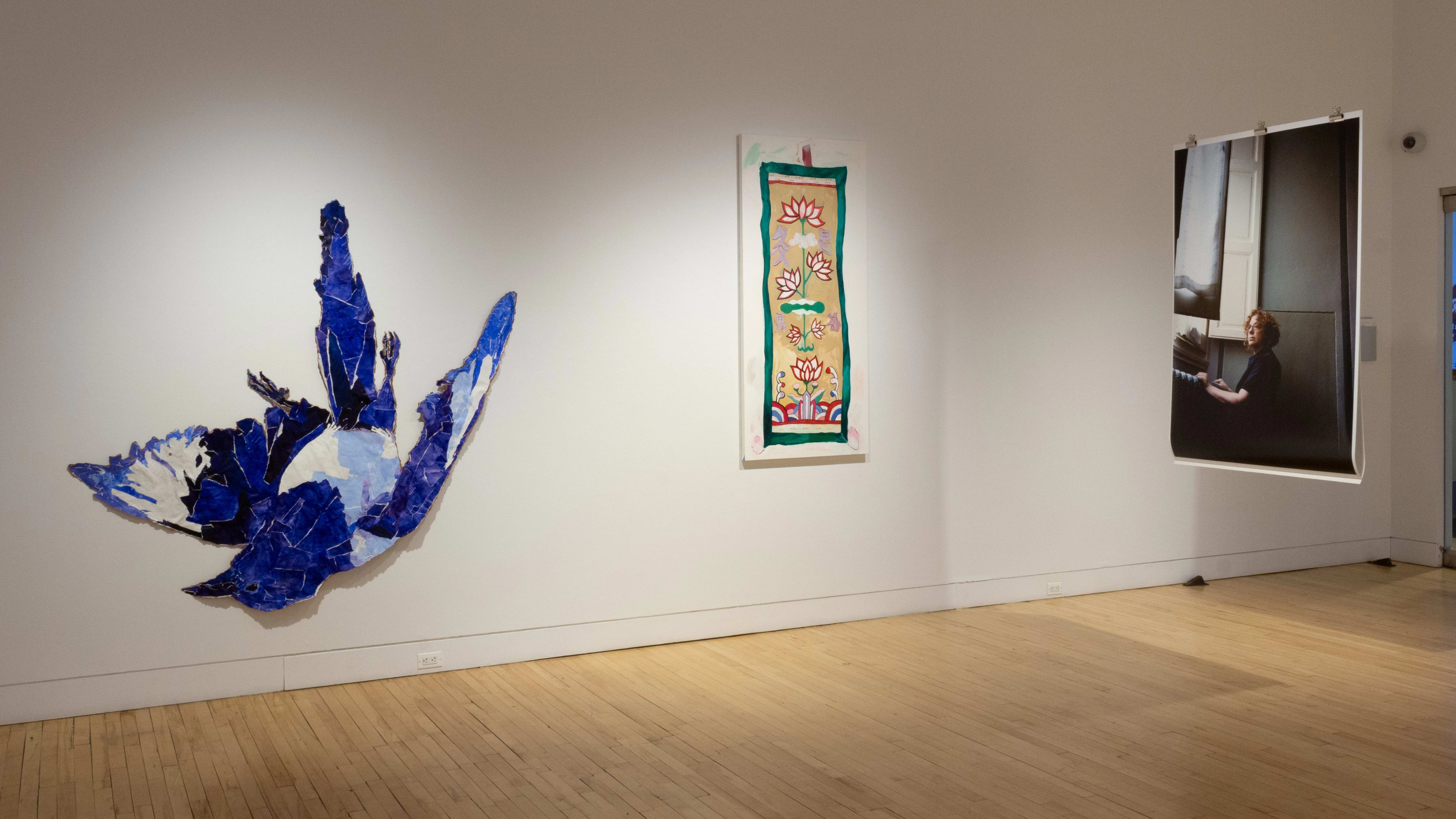 From the other side, the hanging print shows a different photograph. This photo is one of a woman standing near a window. On the wall to the left, there is a painting of a floral pattern, and further, a paper collage of a blue bird falling downward.