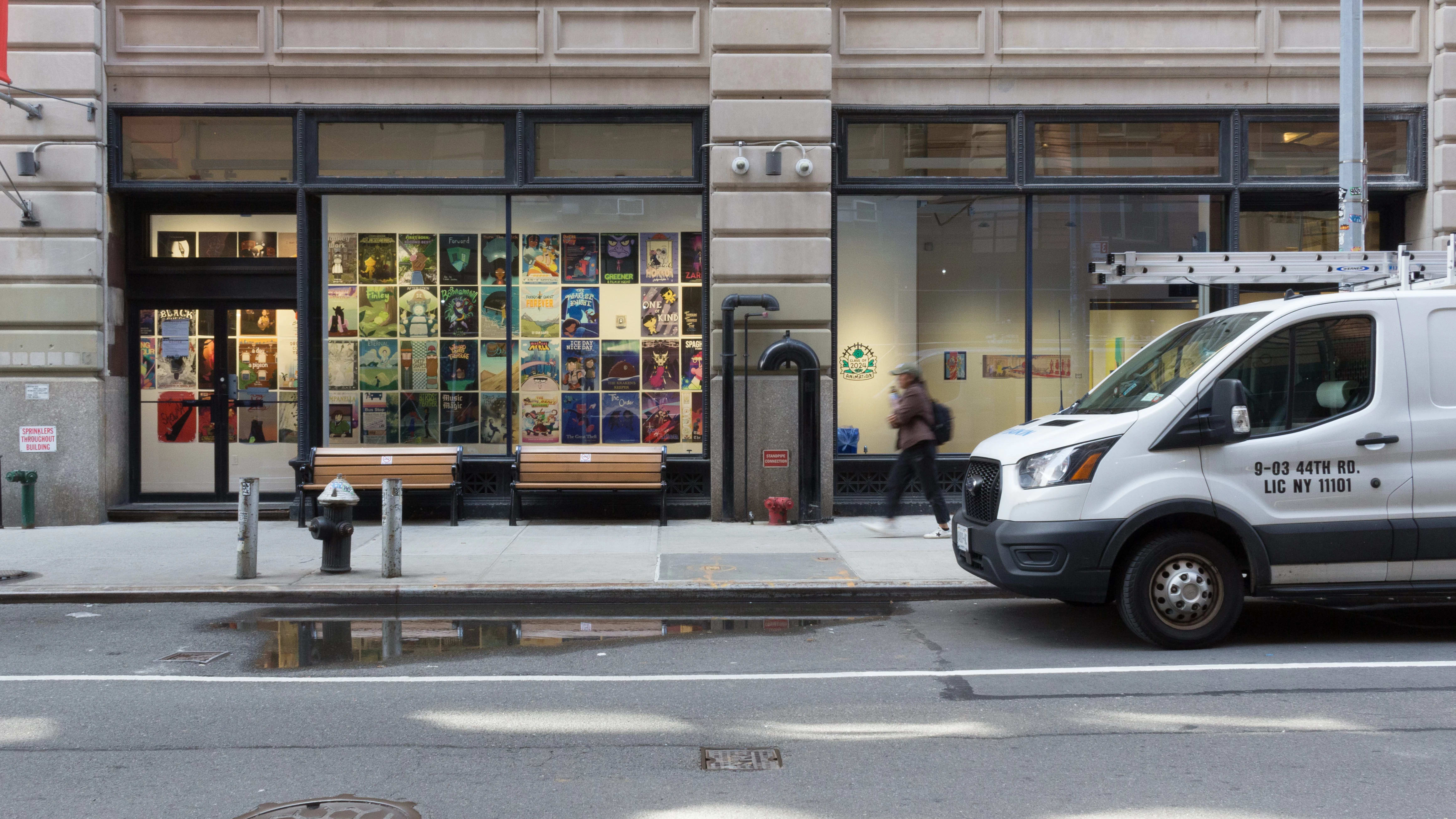 From the street, looking into the windows at the SVA Flatiron Gallery, many posters created by SVA’s BFA Animation Class of 2024 are visible. A person walks by the windows on the sidewalk.
