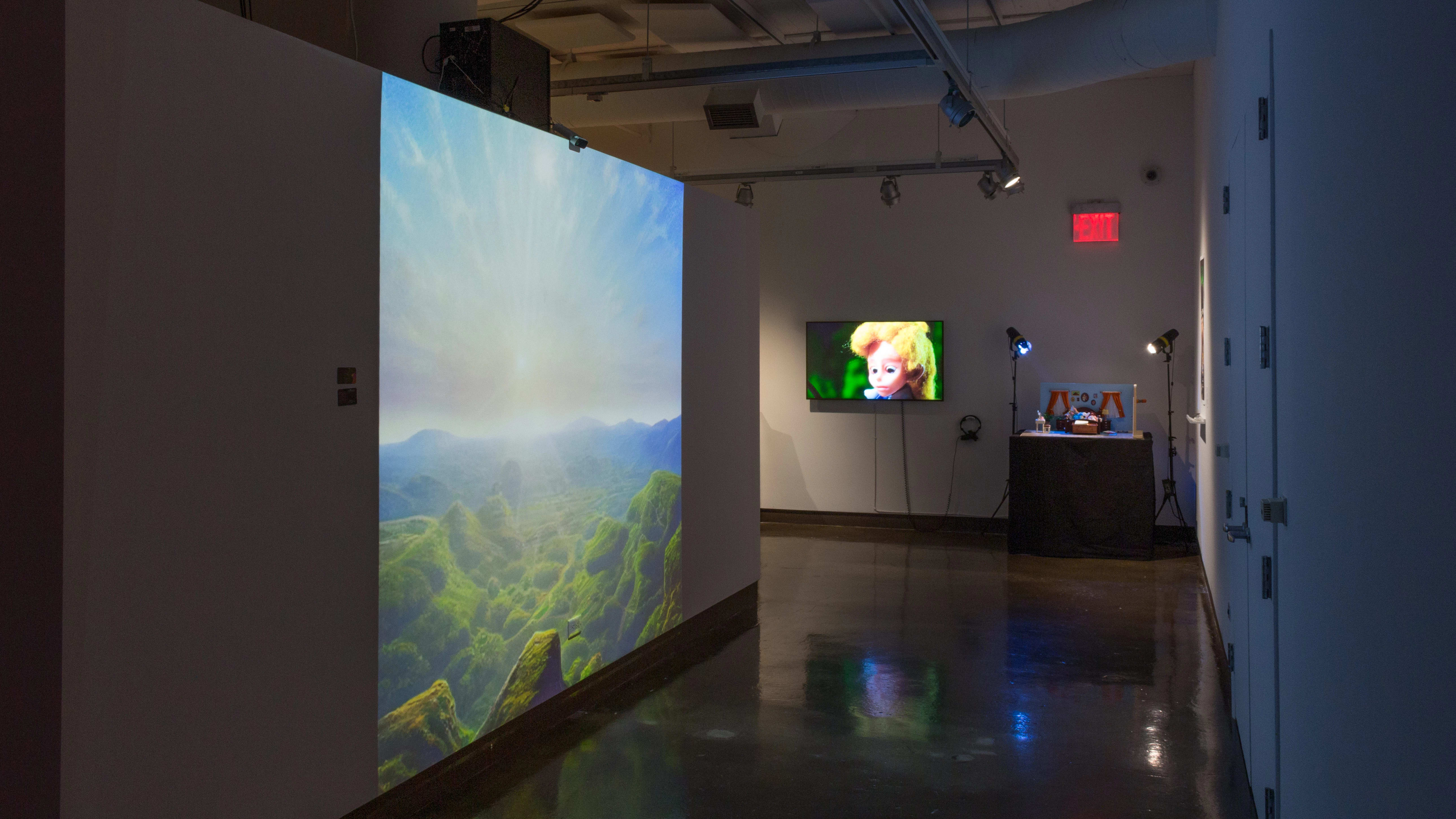 Installation image showing a large projection of a valley to the left, with a monitor and sculptural installation with stand lights in the background.