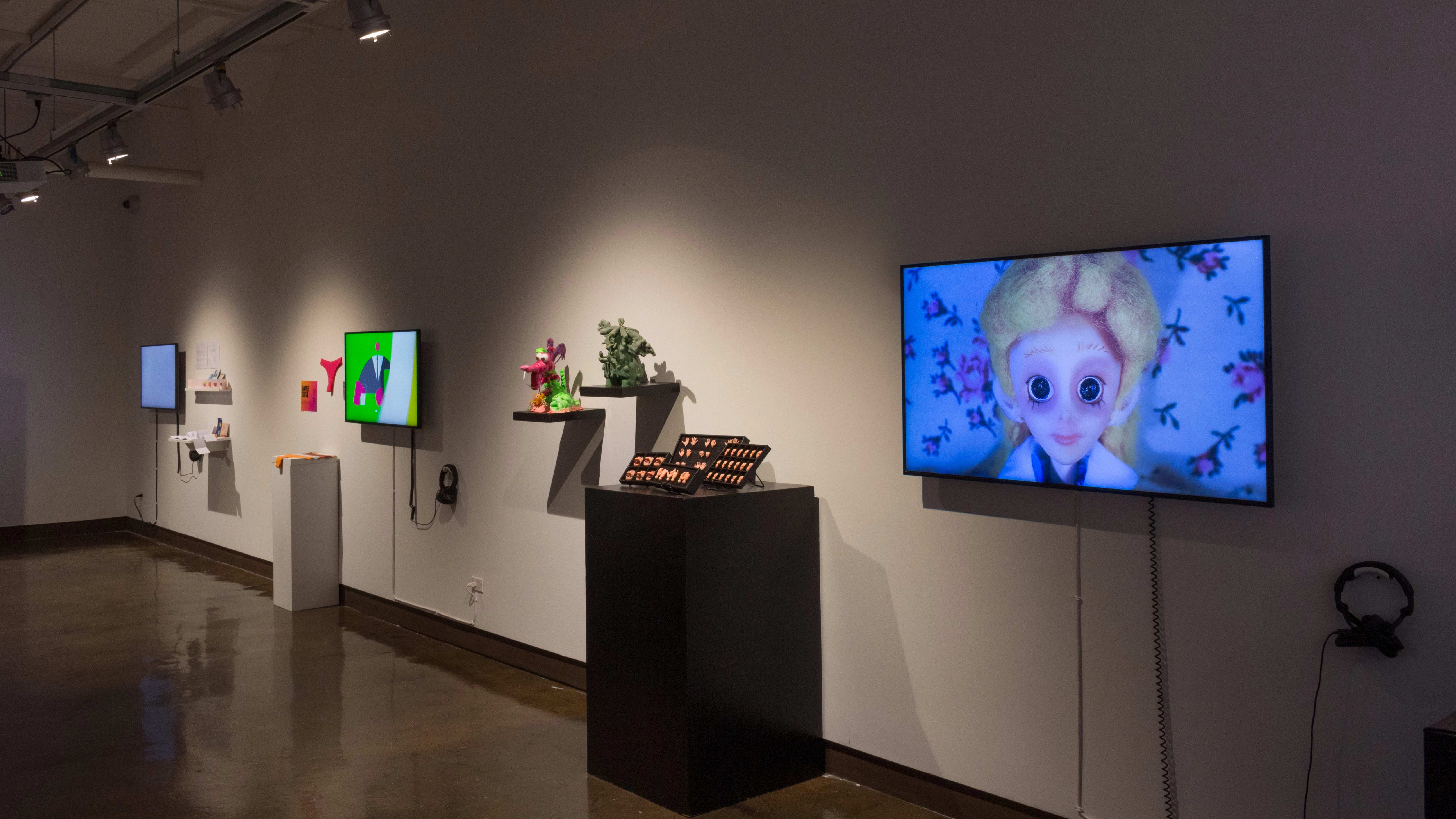 Angled image of a long wall with several monitors, pedestals and shelves with various installations and animated videos.