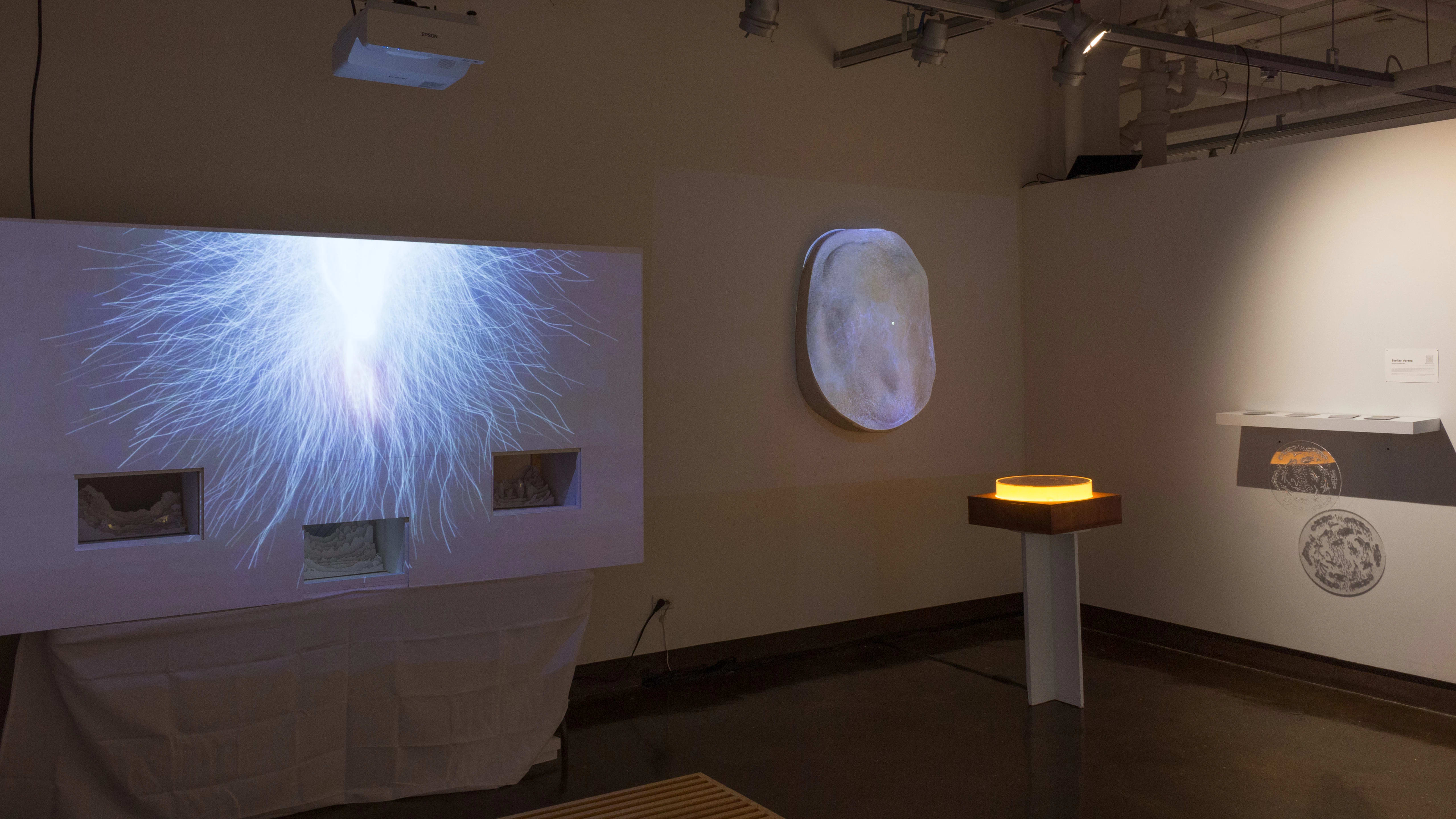 Installation image of an alcove with a large projection of a wavy burst on a false wall with cutouts to the left, and a pedestal and rock projection in the background.